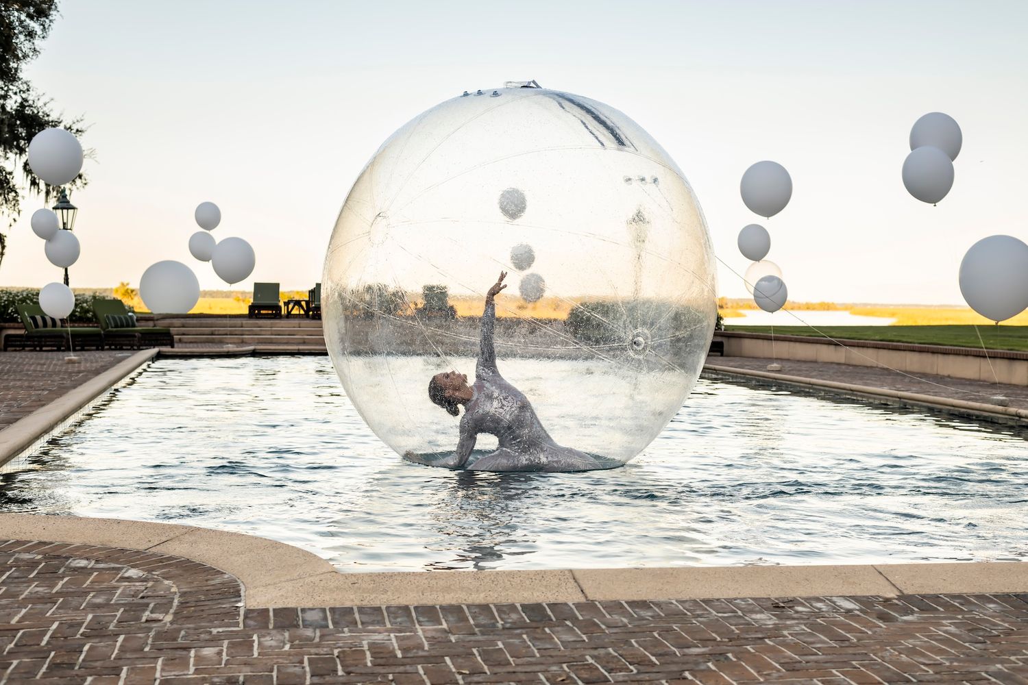A performer in a transparent sphere floats on water with gray balloons floating in the sunset sky background.