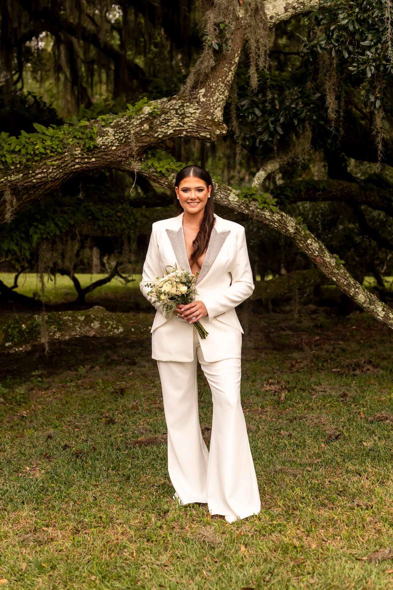 A white pantsuit and bouquet against a backdrop of moss-draped trees in a dreamy outdoor setting.