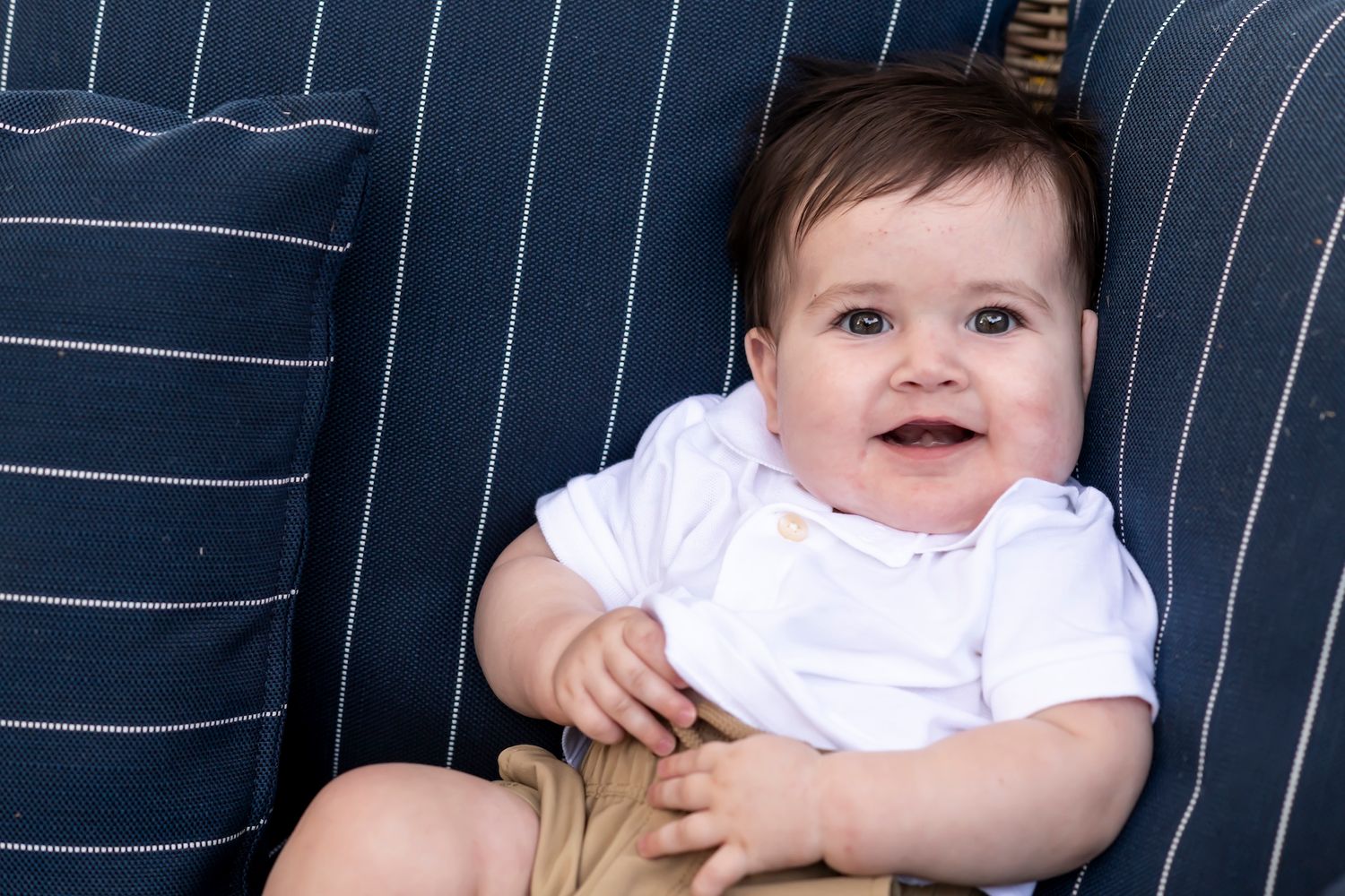 A smiling baby wearing a white top sits on a navy blue striped couch cushion with a joyful expression.