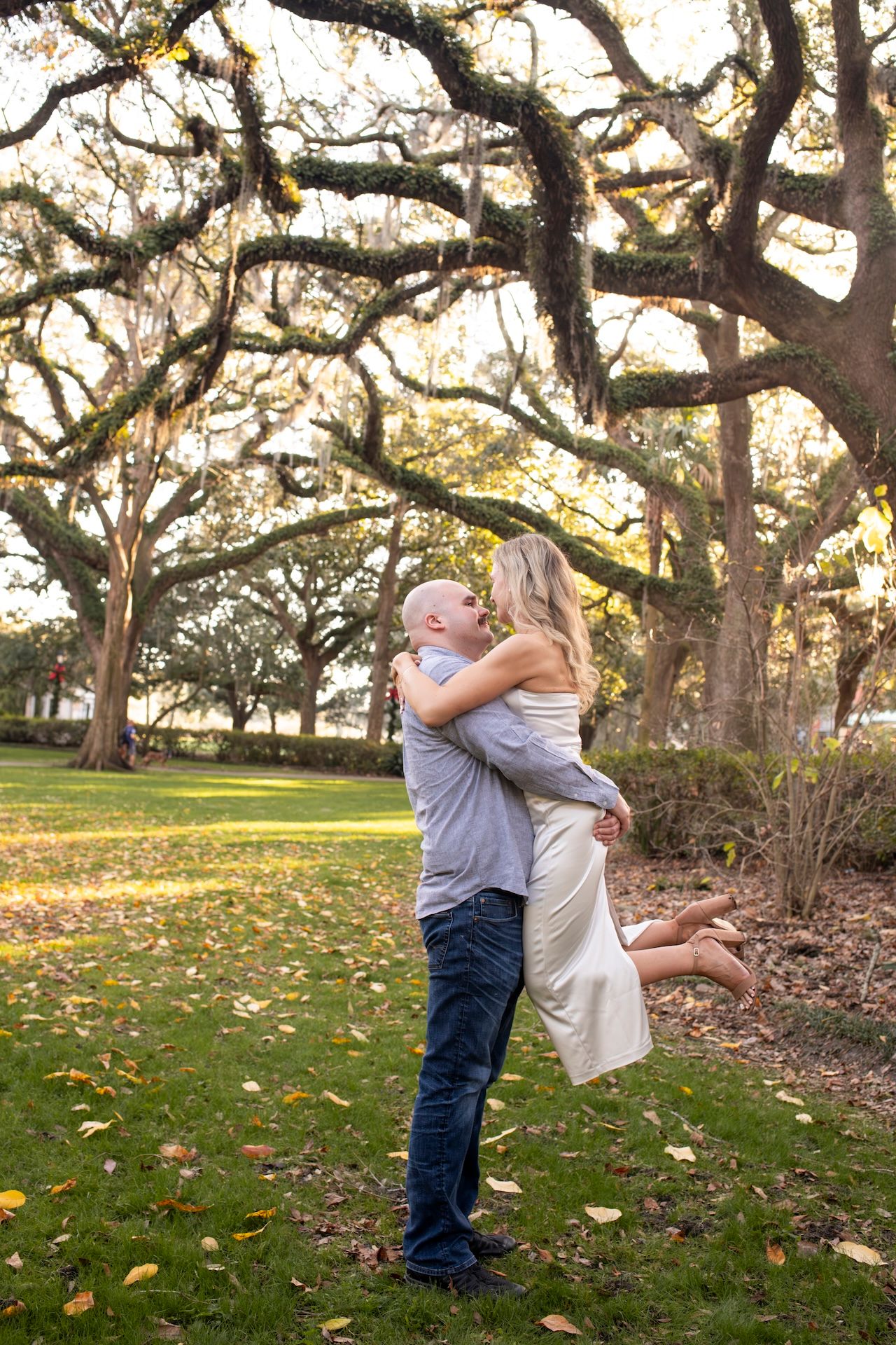 A romantic couple embraces under moss-draped oak trees in a sunlit park with fallen autumn leaves on the ground.