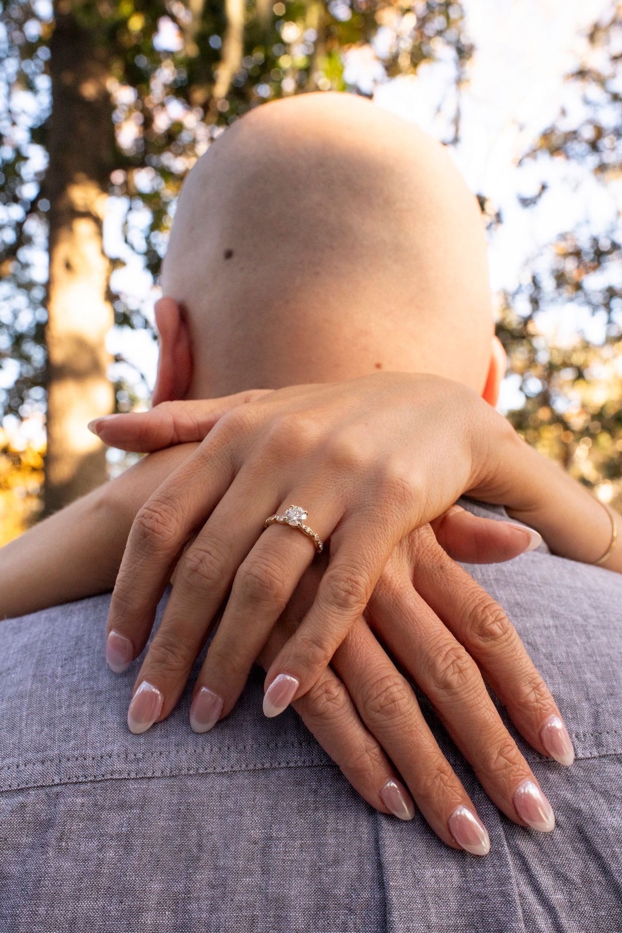 Close-up of hands with French manicure and engagement ring resting on grey fabric with sunlit trees in background.