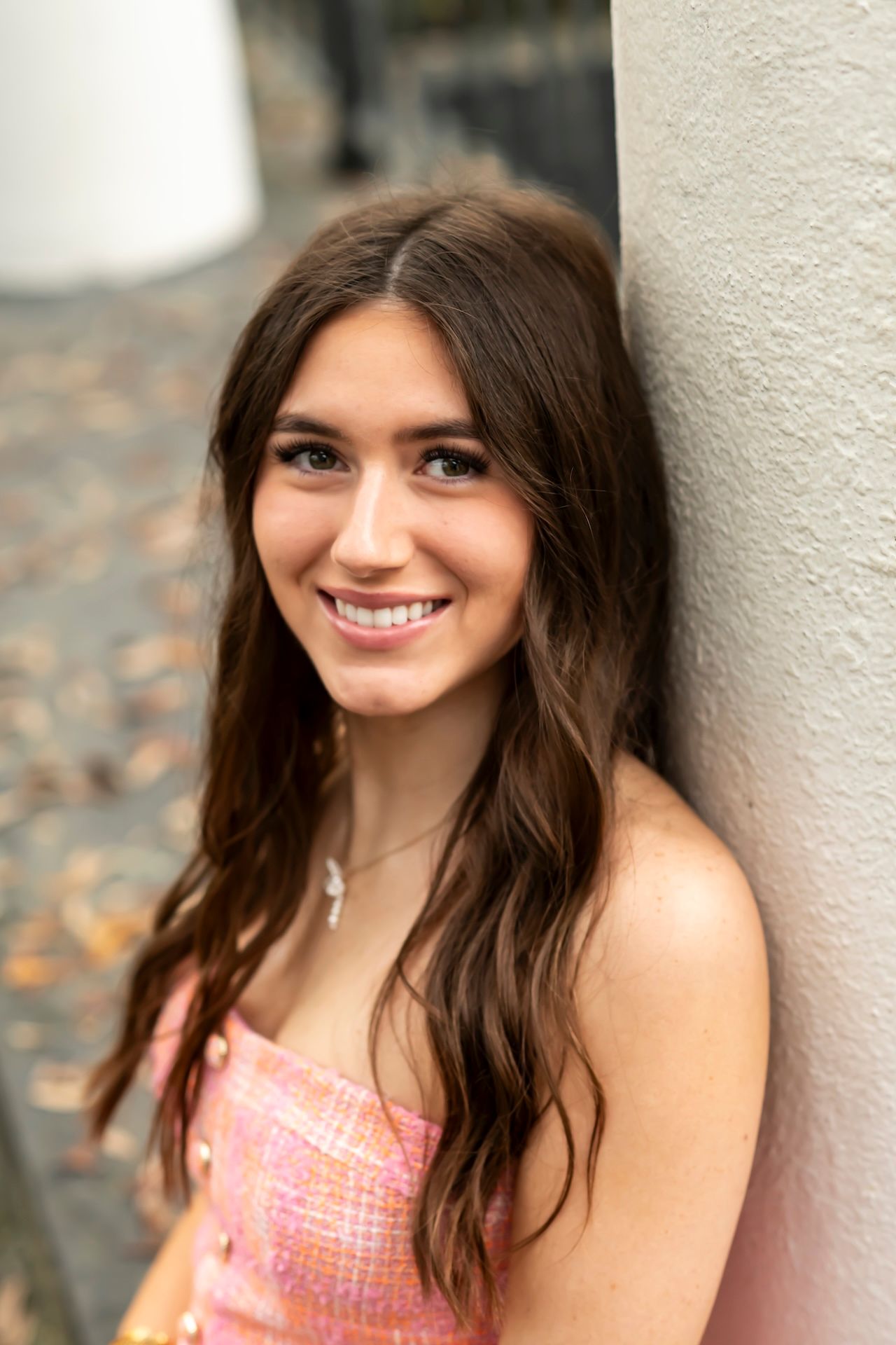 A smiling person wearing a pink patterned top leans against a light colored wall in an outdoor portrait setting.