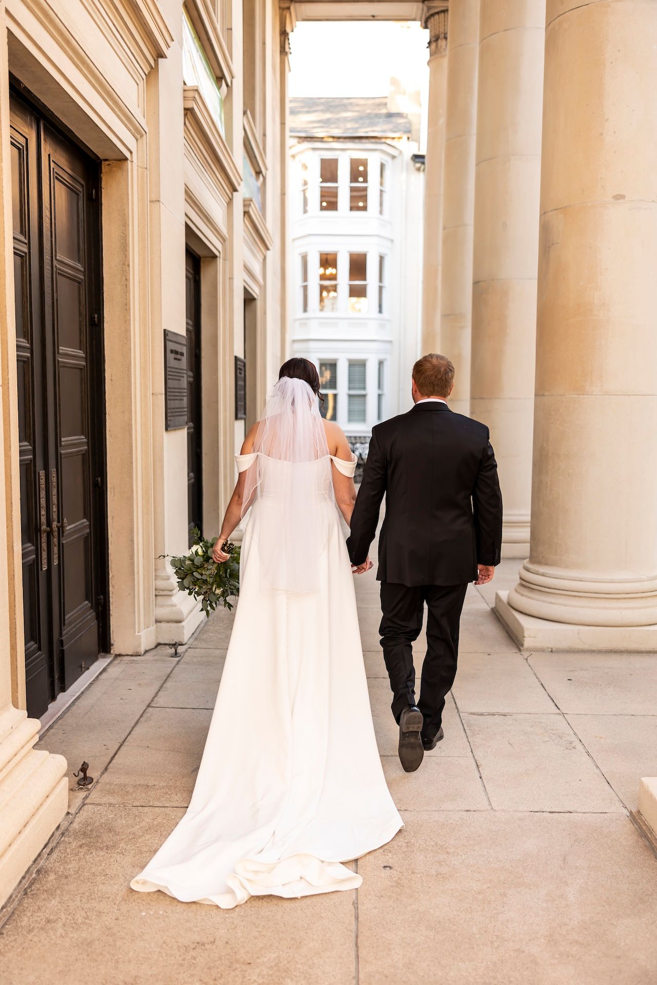 A bride and groom walk hand in hand down a columned walkway, her long white dress trailing behind them.