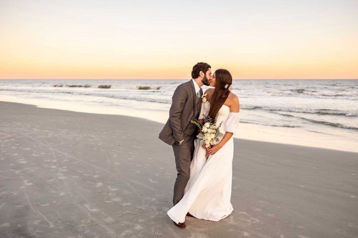 A couple shares a romantic moment on a beach at sunset while holding a wedding bouquet, with waves crashing in the background.