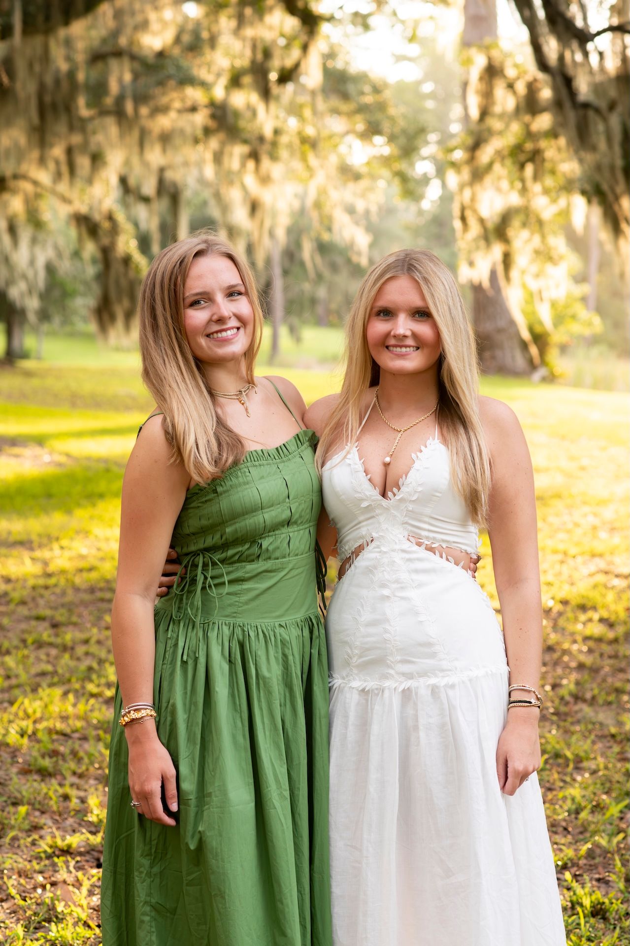 Two smiling blonde women pose outdoors in green and white maxi dresses under Spanish moss-draped trees.