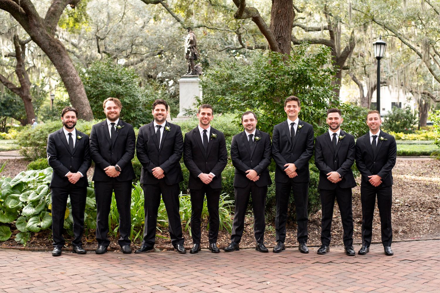A group of groomsmen in matching black suits and ties stand in a line at a garden setting with trees and lamp posts.