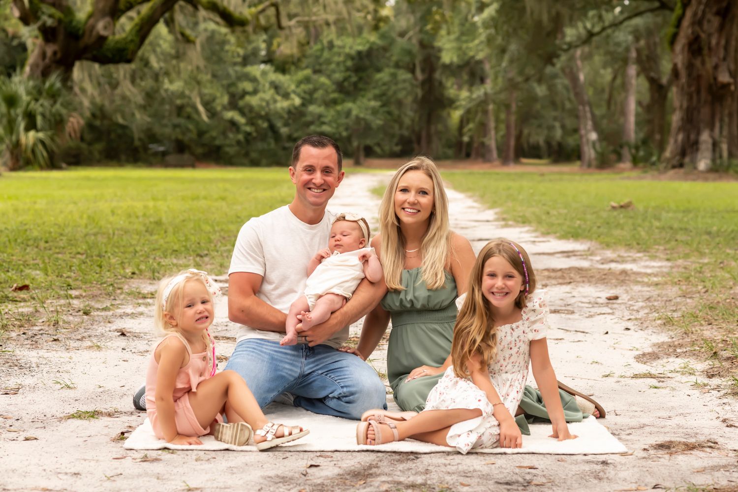 A family sits together on a blanket for an outdoor portrait session along a sandy path surrounded by oak trees.