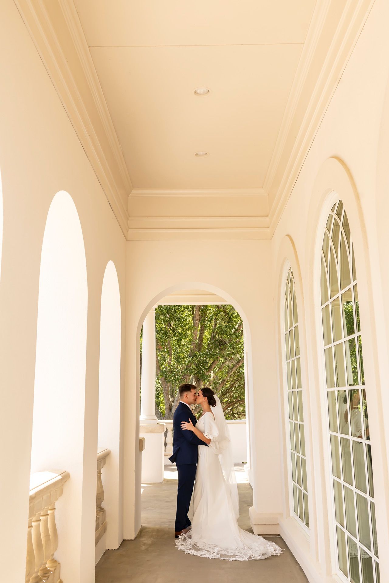 A couple shares an intimate moment in an elegant white colonnade walkway with arched windows and natural lighting.