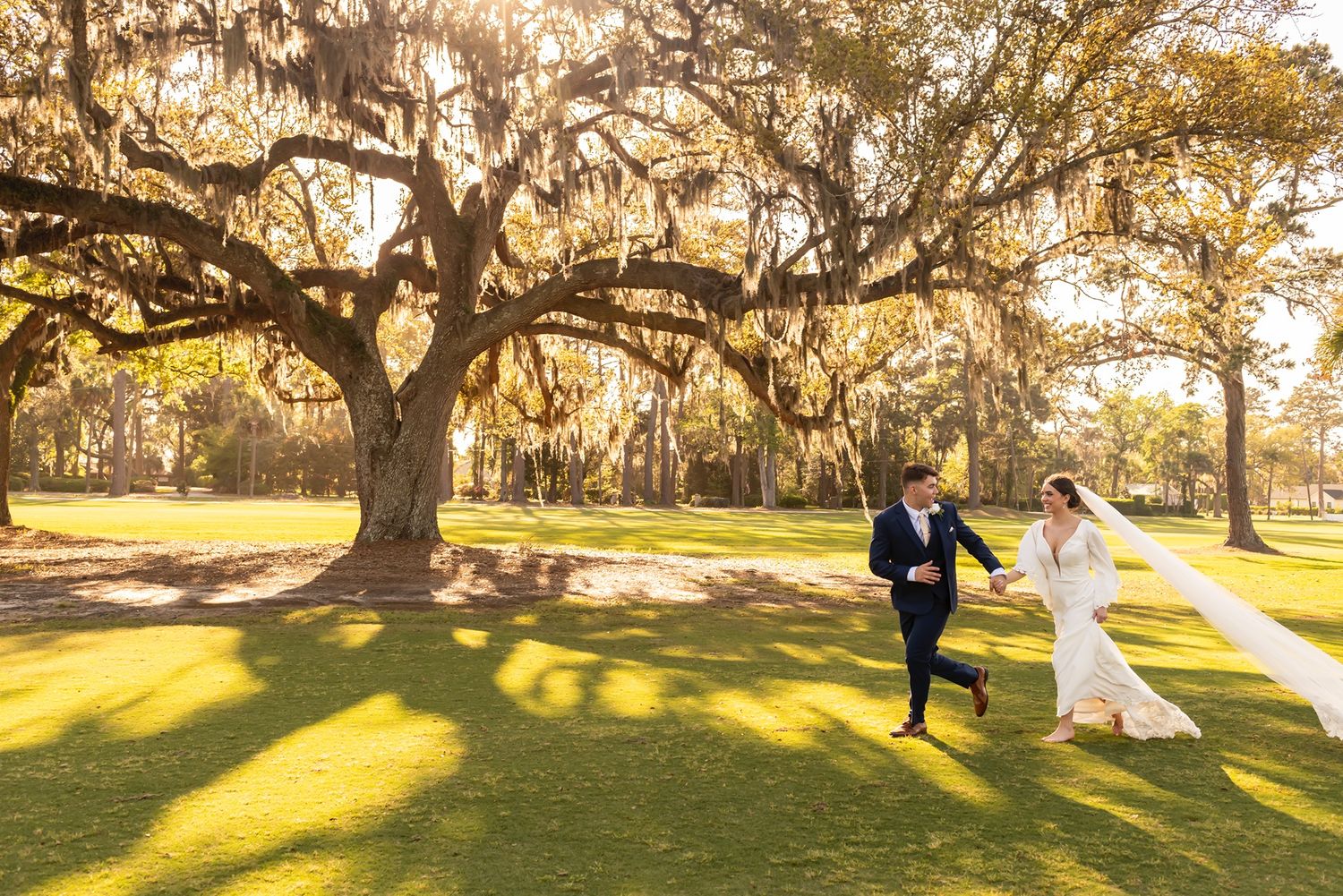 A playful wedding couple runs beneath sprawling oak trees with golden sunlight streaming through Spanish moss.