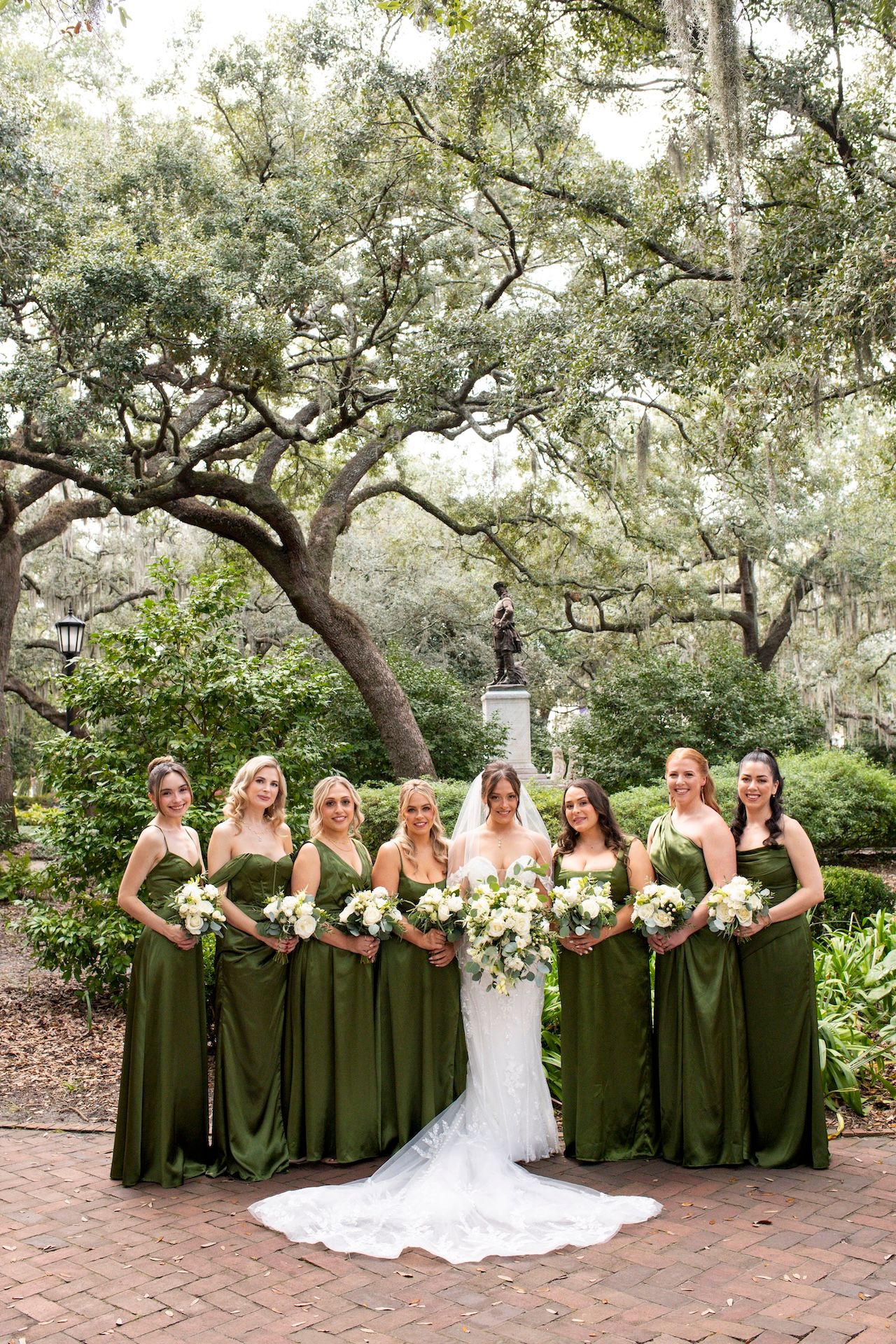 Wedding party wearing olive green bridesmaid dresses poses together in a garden setting with brick pathways.