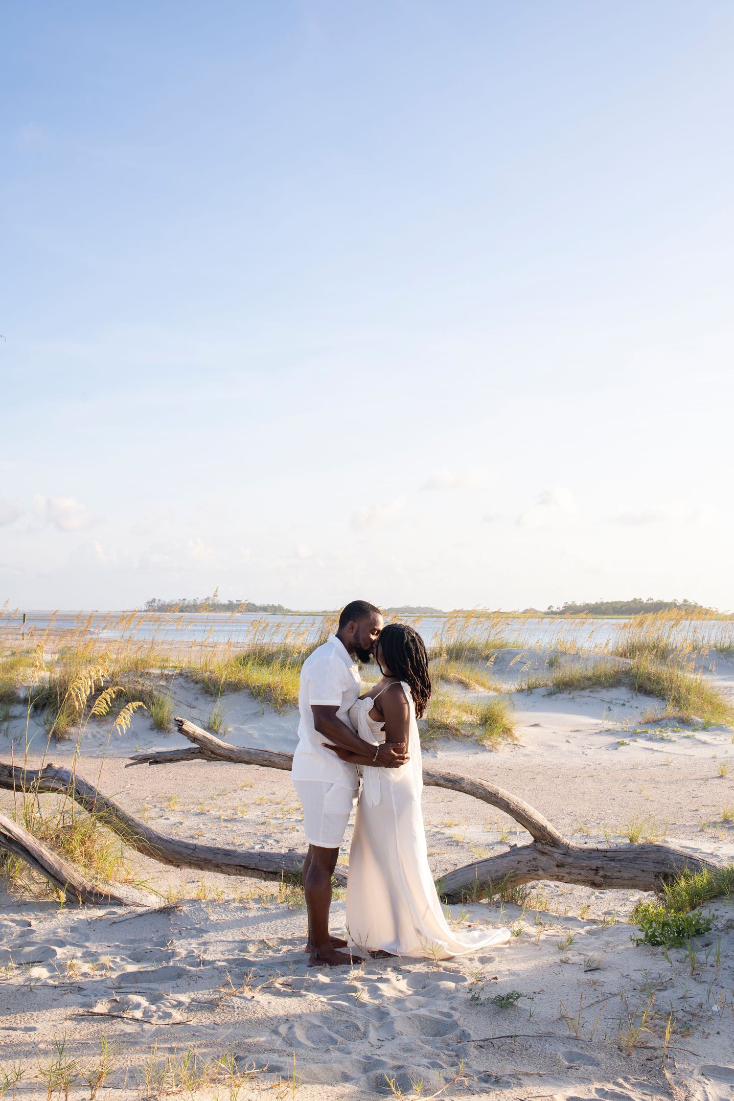A romantic couple embraces on a sandy beach with sea grass and coastal waters in the background during golden hour.