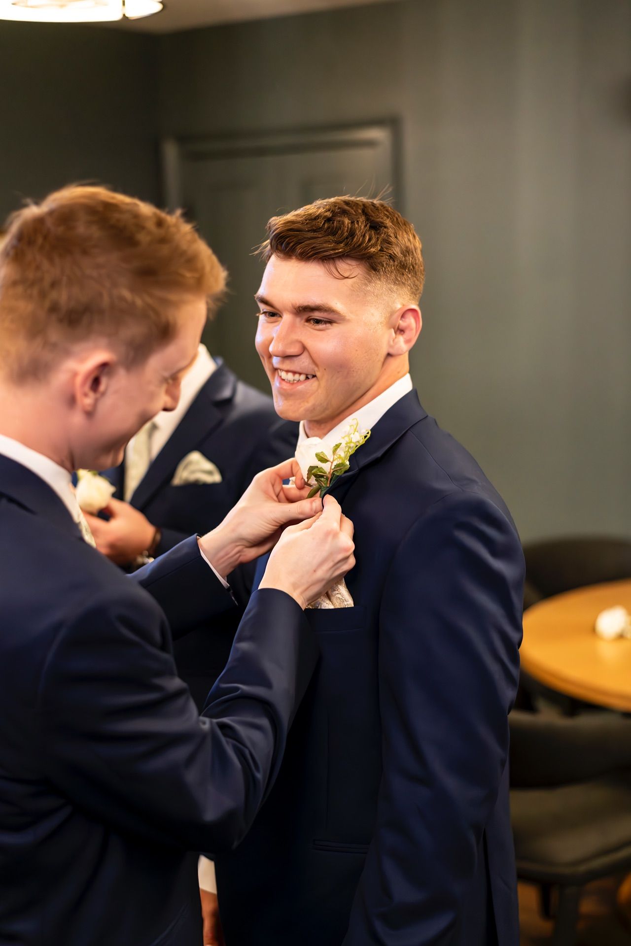 A groomsman helps adjust another's boutonniere at a formal wedding preparation in navy suits.