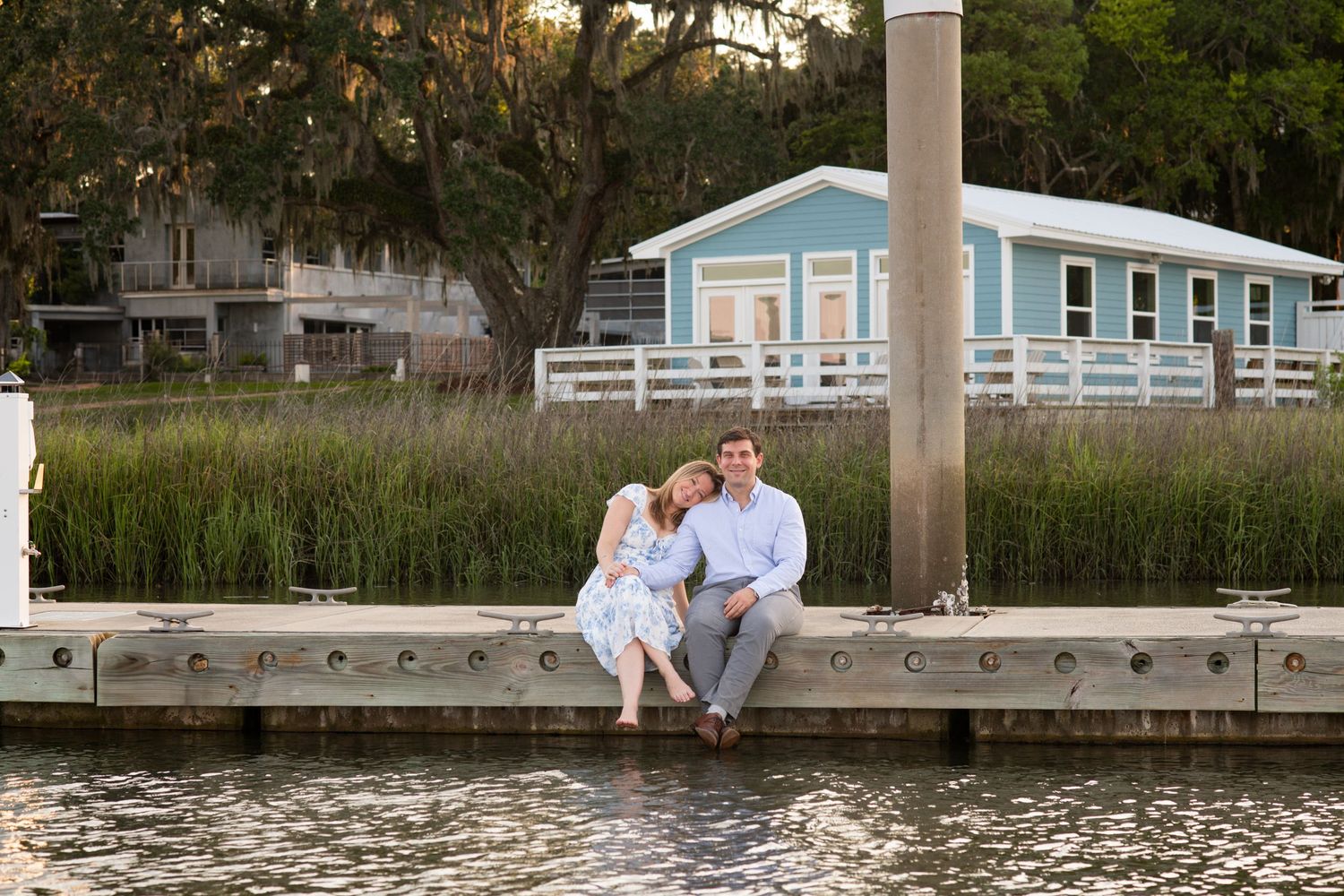 A couple sits together on a wooden dock by a waterfront with a light blue cottage and marsh grass in the background.
