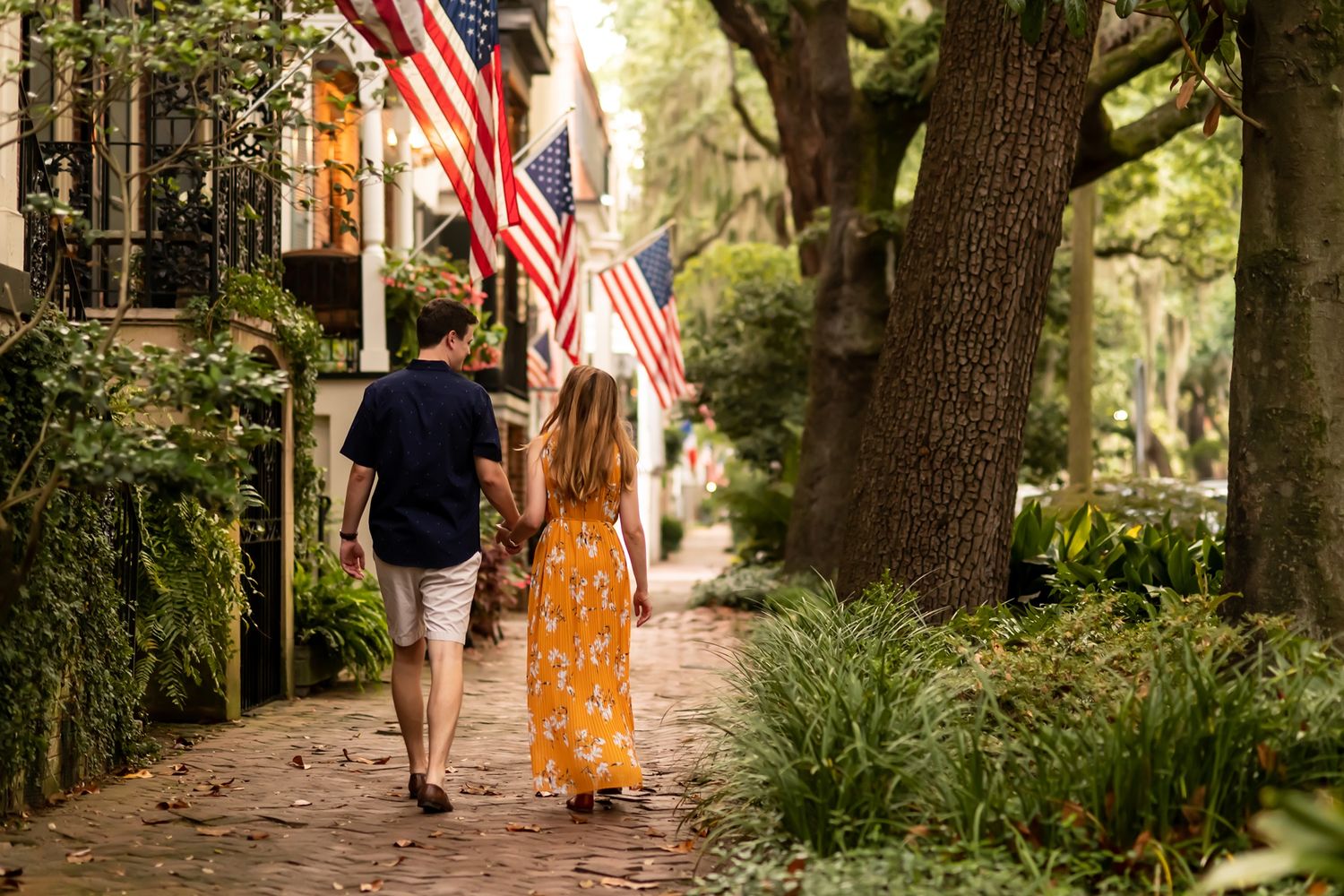Two people walking together on a shaded brick pathway with American flags and lush greenery in historic Charleston.