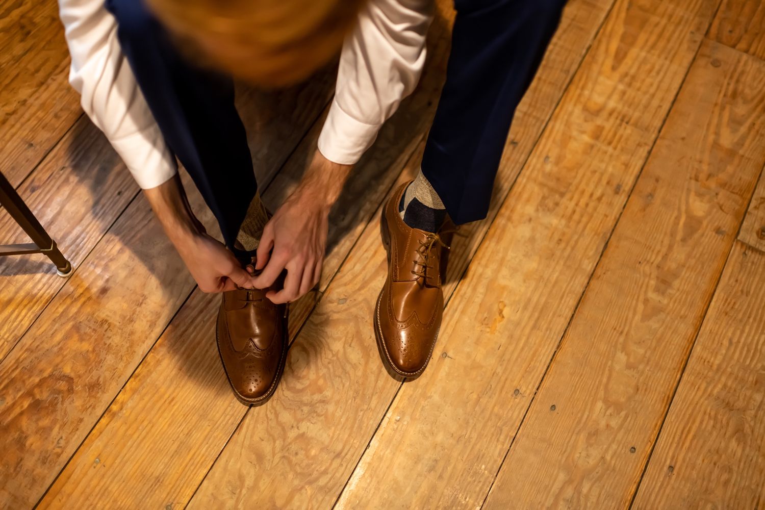 Someone ties laces on brown leather dress shoes while standing on a wooden floor.