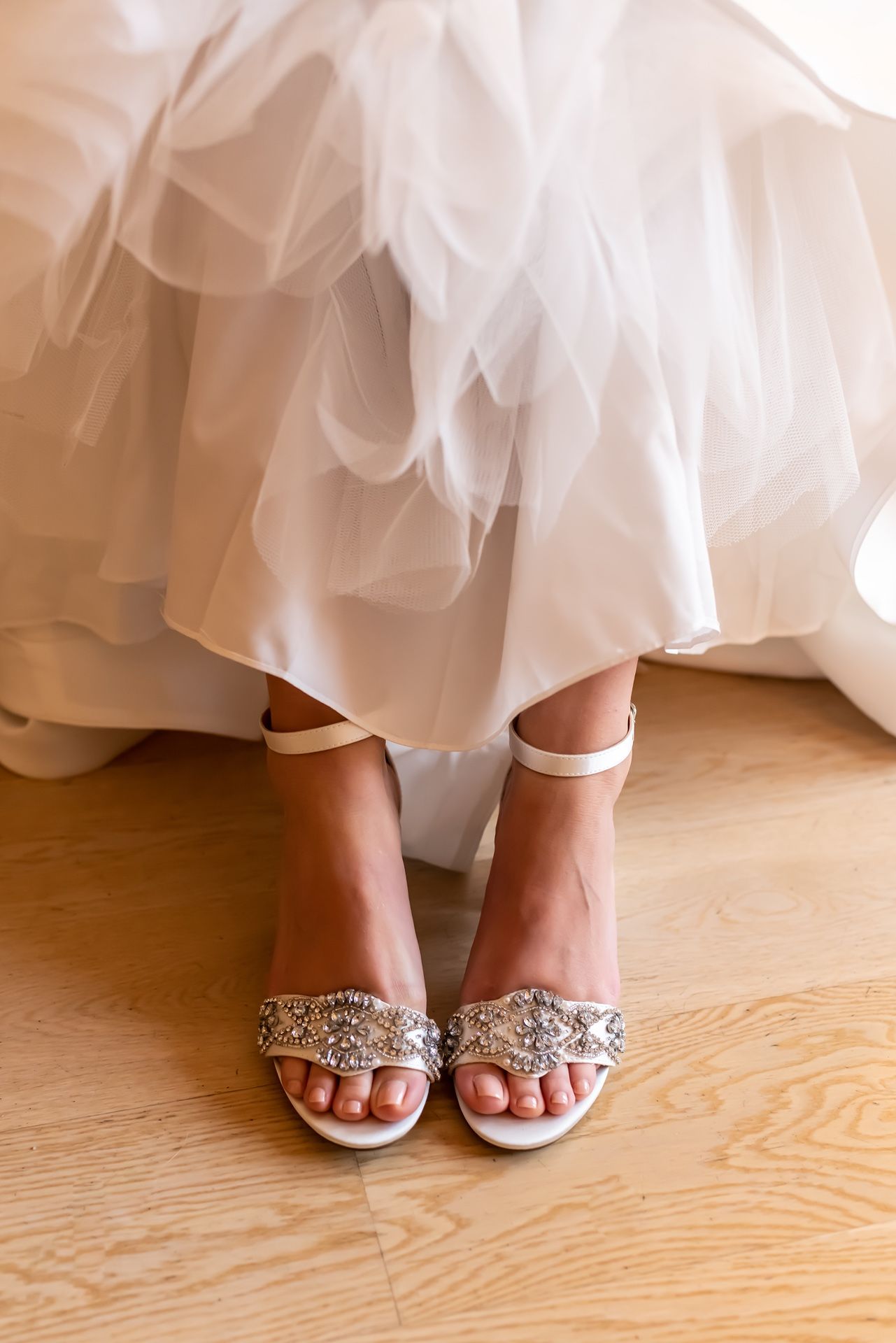 A close-up view of elegant bridal sandals with crystal embellishments peeking out from under a flowing white wedding gown.