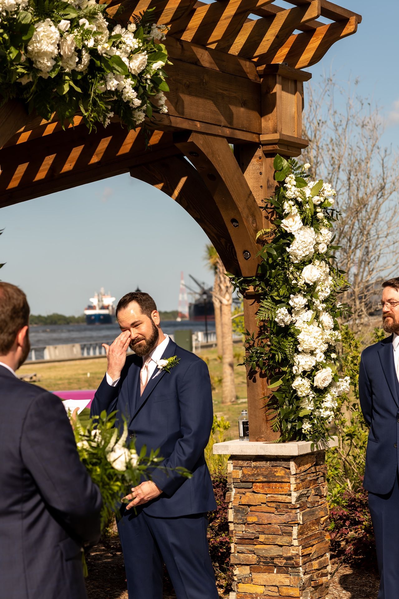 A groom wipes away tears during an emotional moment under a wooden pergola decorated with white flowers at an outdoor wedding.