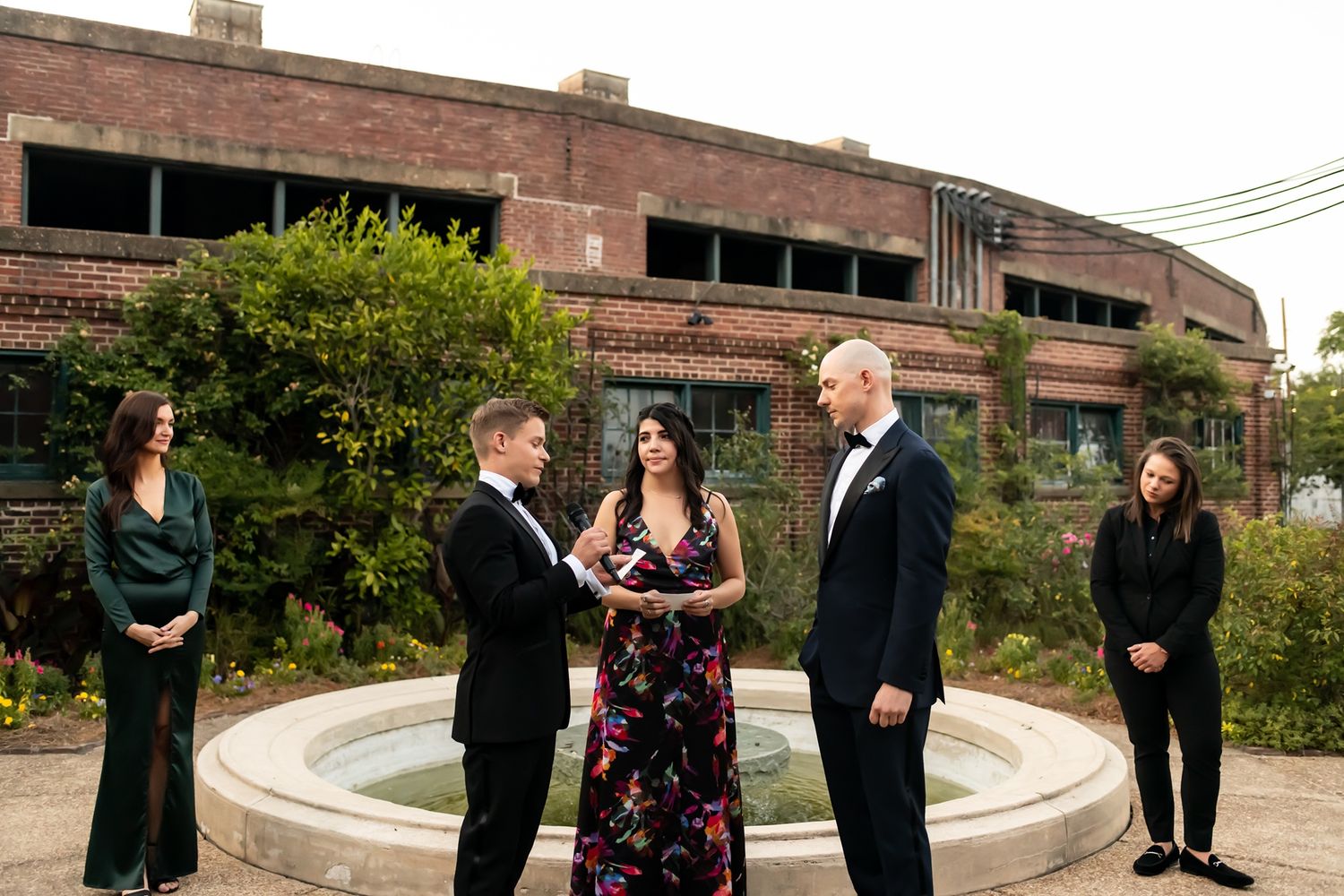 A small group gathers around an outdoor stone fountain with an abandoned brick building in the background.