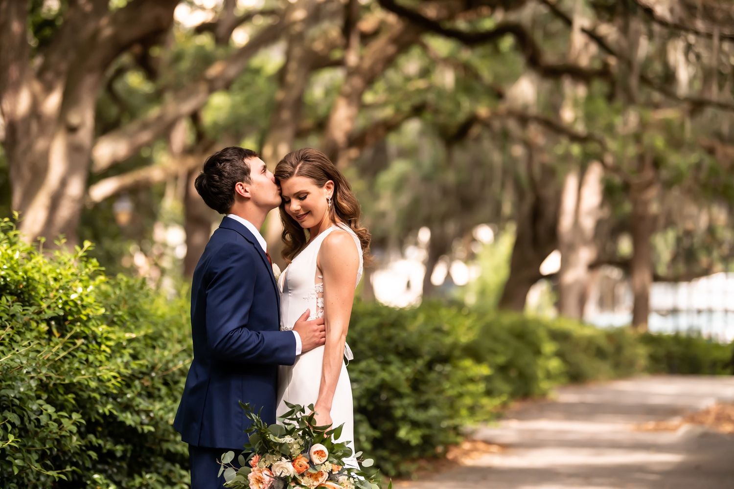 Bride and groom share romantic moment on tree-lined path during outdoor garden wedding photoshoot.