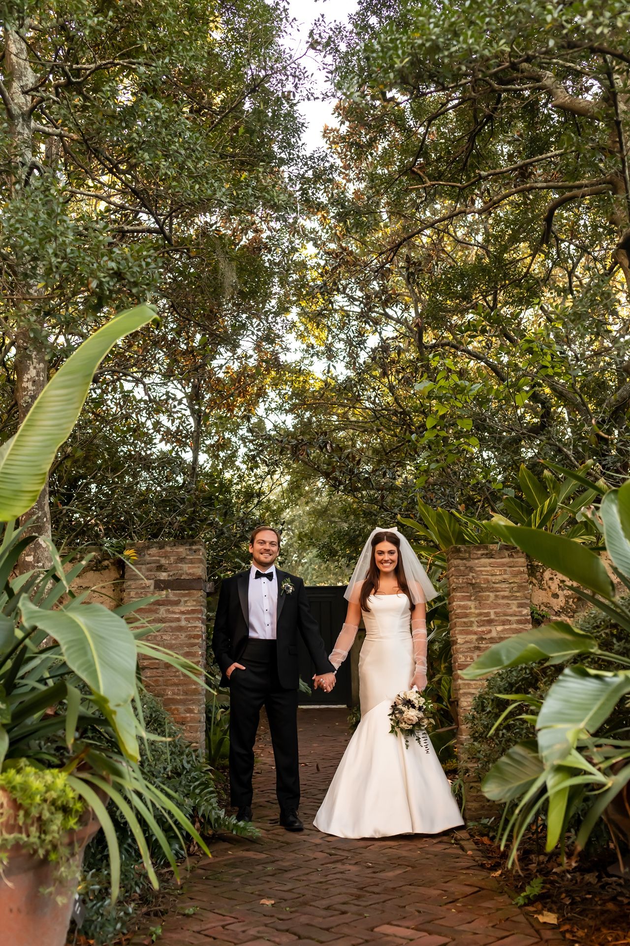 A happy couple stands in a lush garden walkway framed by brick walls and tropical plants on their wedding day.