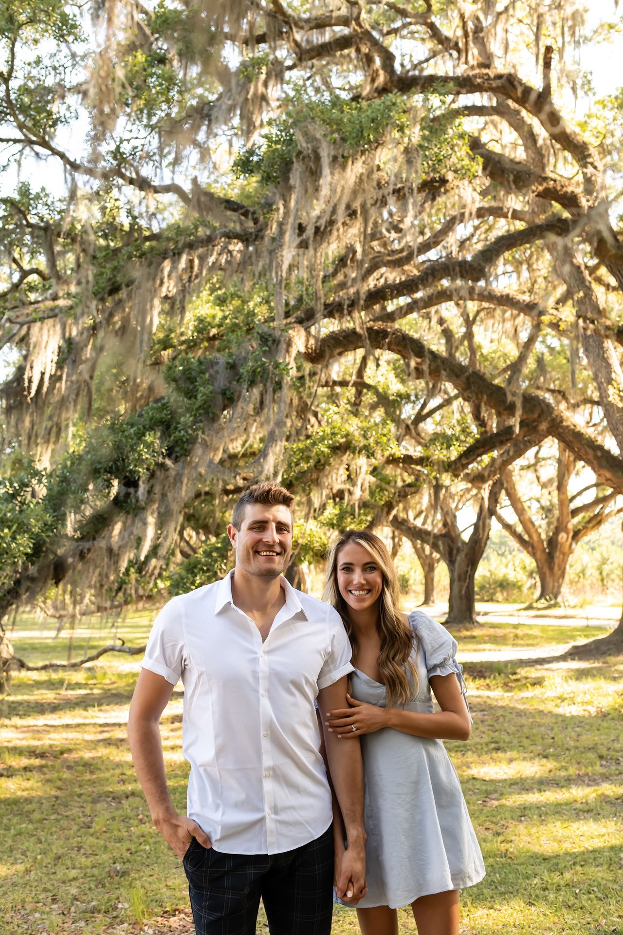 Two people stand together under majestic Spanish moss-draped oak trees in a sun-dappled Southern setting.