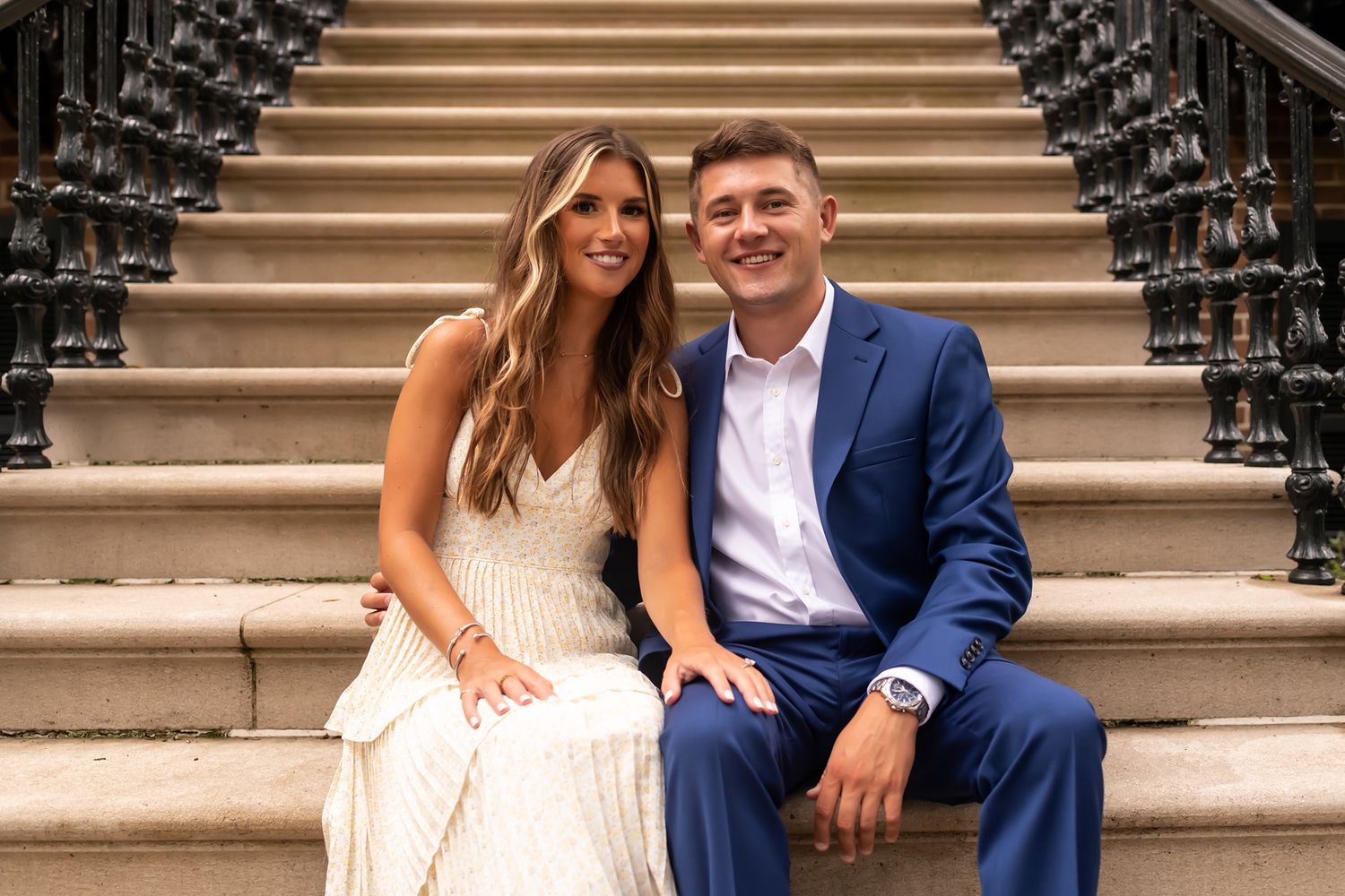 A couple sits together on elegant brownstone stairs, wearing formal attire with her in white and him in navy.