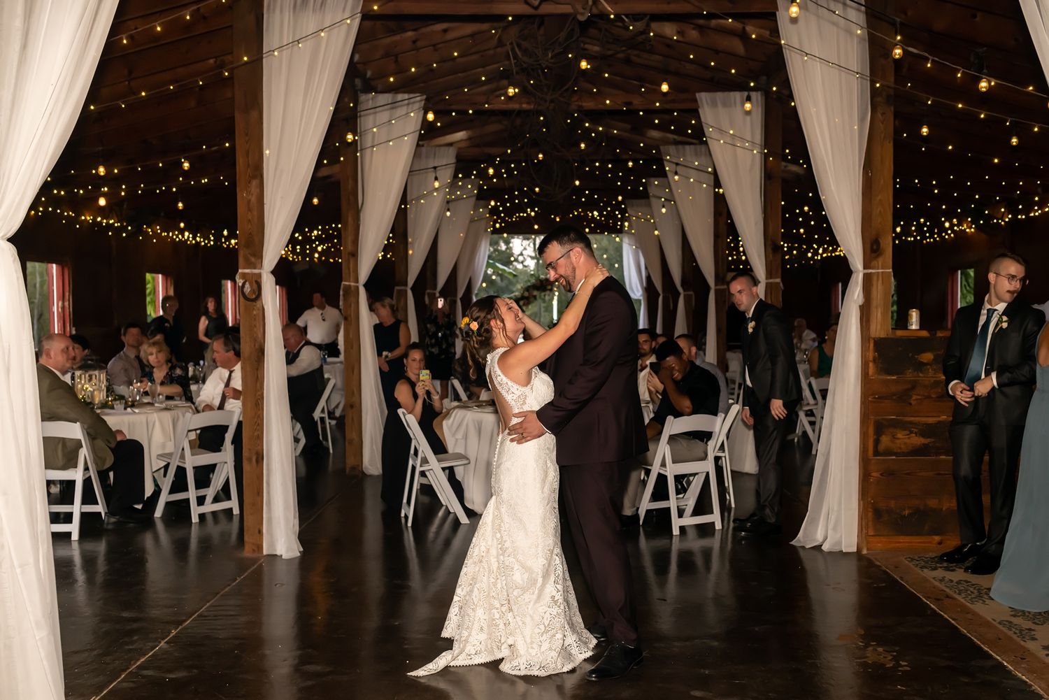 Couples dance during an elegant evening wedding reception in a rustic barn venue decorated with white drapes and twinkling lights.