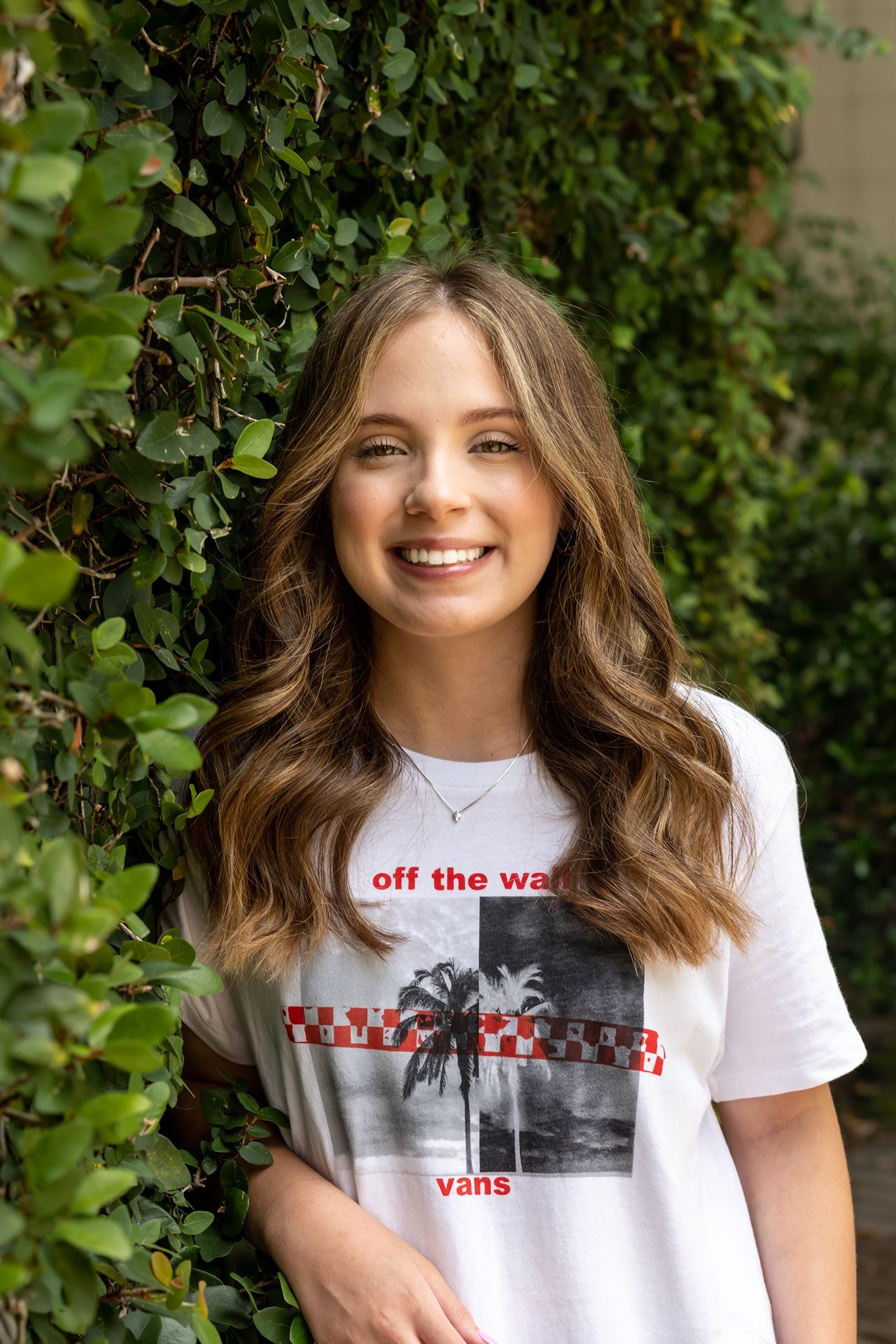 Person smiling outdoors wearing a white Vans t-shirt against a green leafy background.