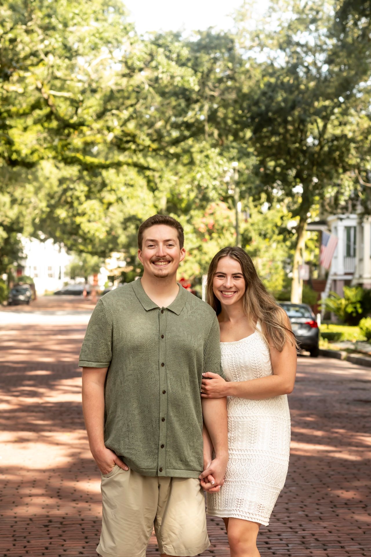 Two people stand close together smiling on a brick walkway surrounded by green trees on a sunny day.