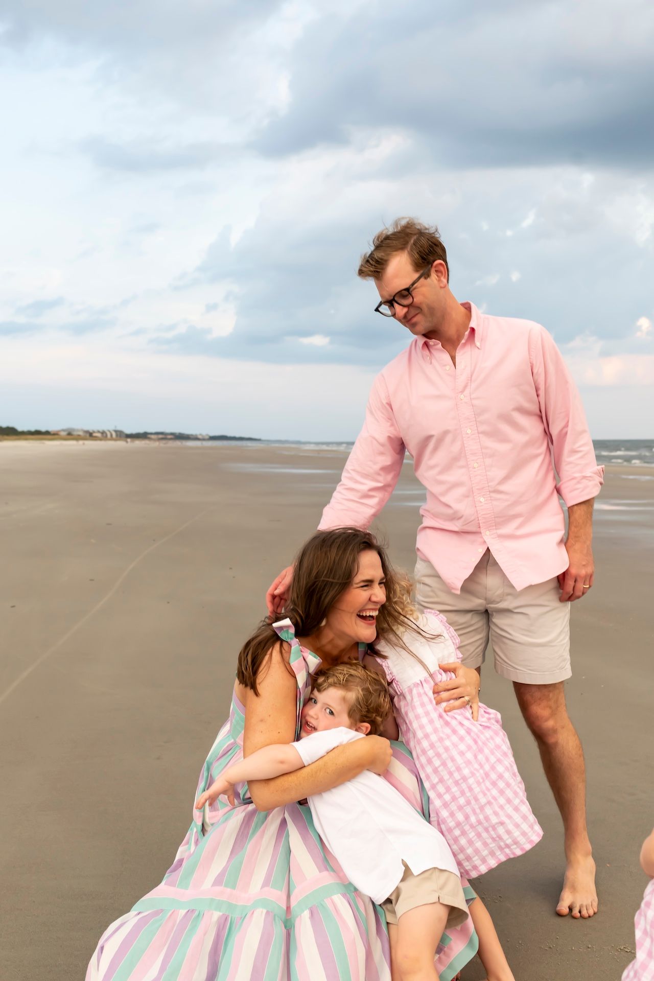 A joyful family moment captured on a wide sandy beach under cloudy skies, wearing coordinated pink and white outfits.