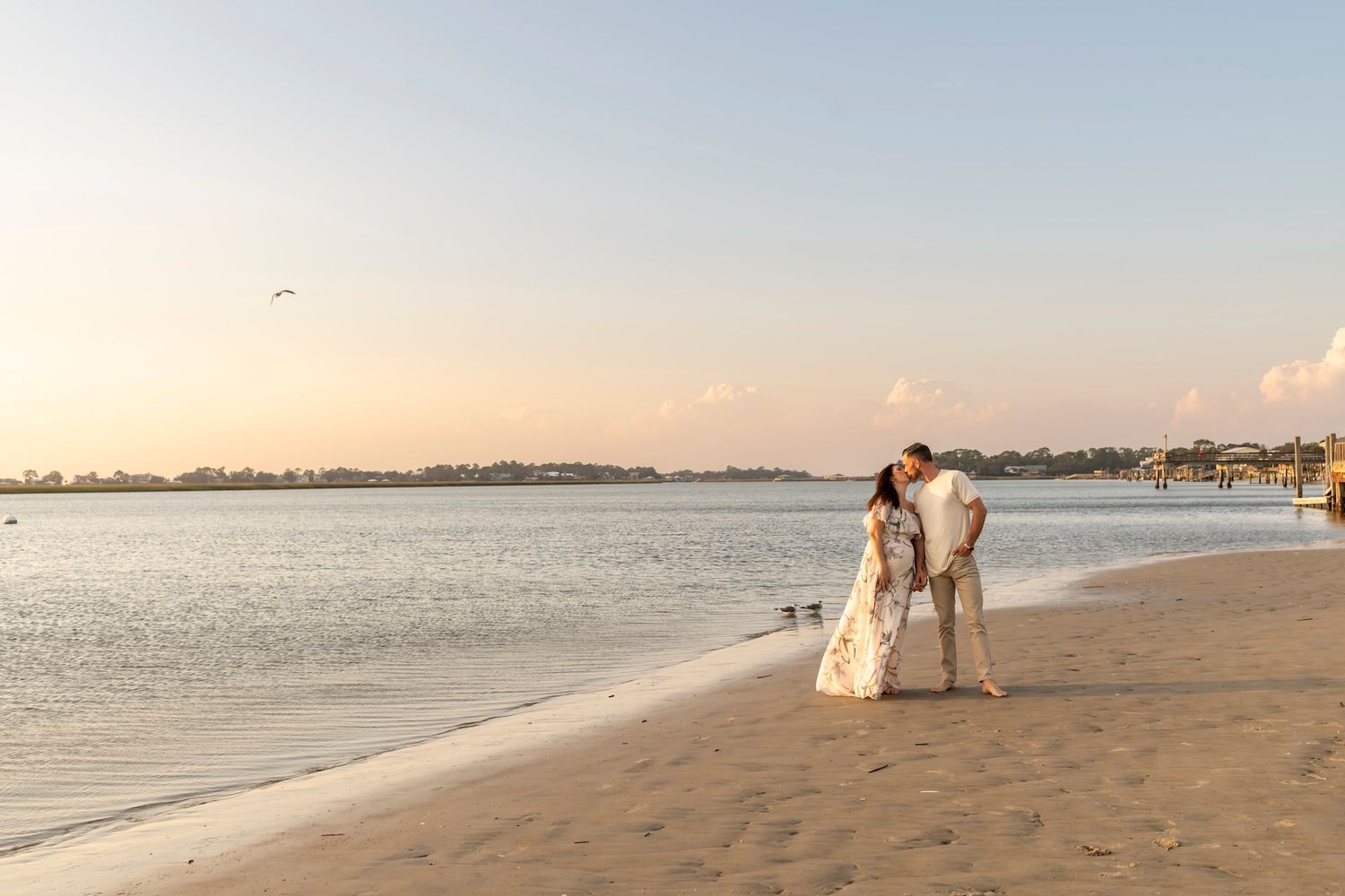 A couple in formal attire stands together at sunset on a sandy beach with calm waters and distant shoreline.
