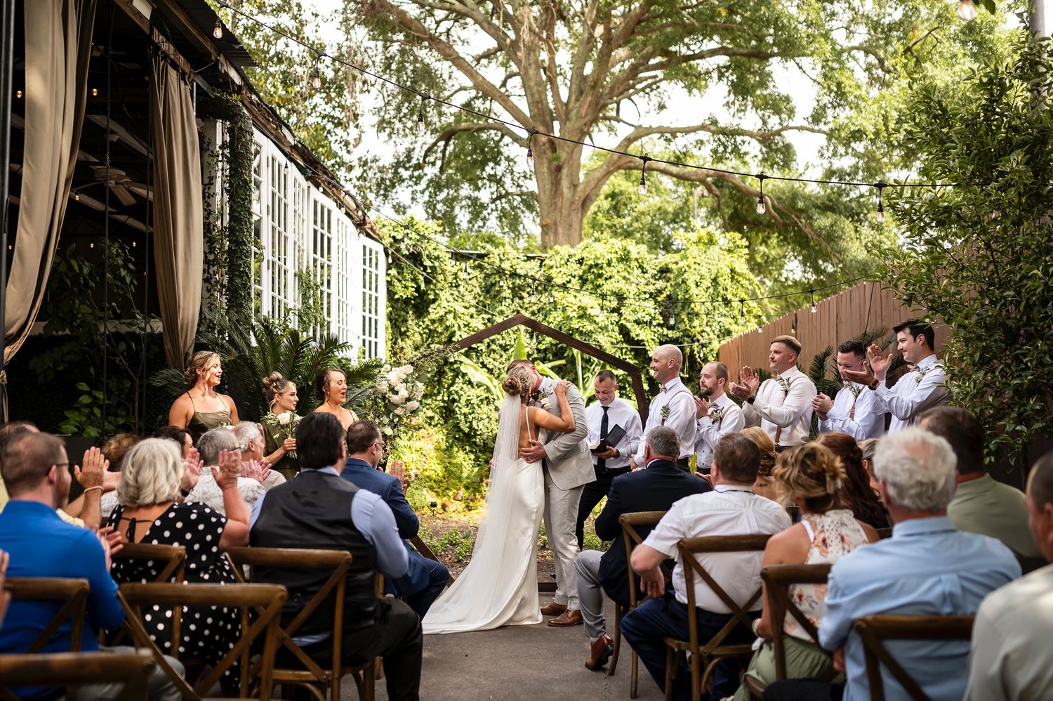 An outdoor garden wedding ceremony with guests seated on wooden benches under a lush tree canopy.