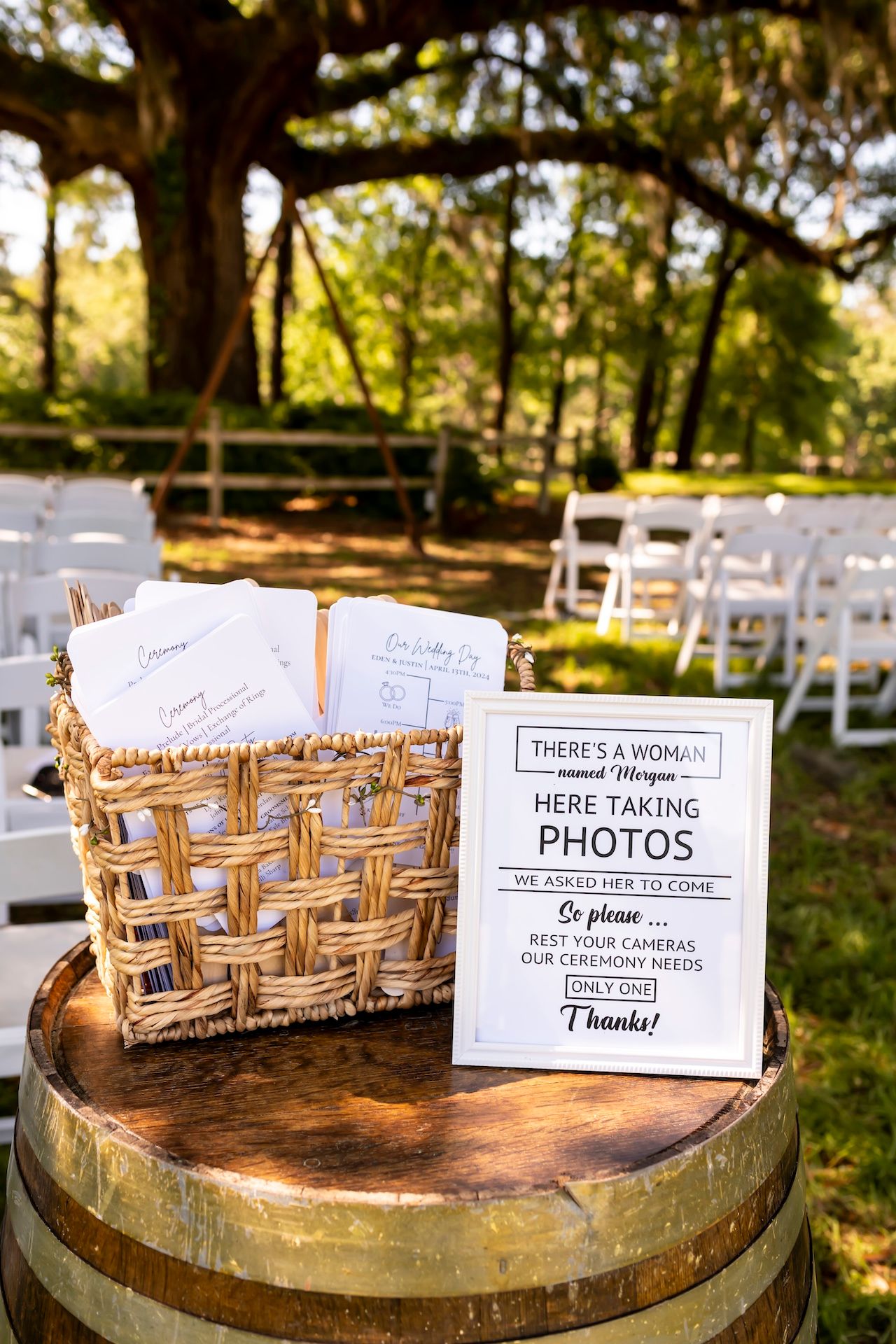 Wedding setup in outdoor garden with white chairs and rustic barrel featuring photography sign and woven basket.