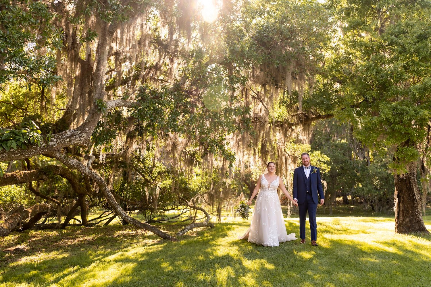 A romantic wedding photo under Spanish moss draped oak trees with golden sunlight streaming through in a scenic garden setting.