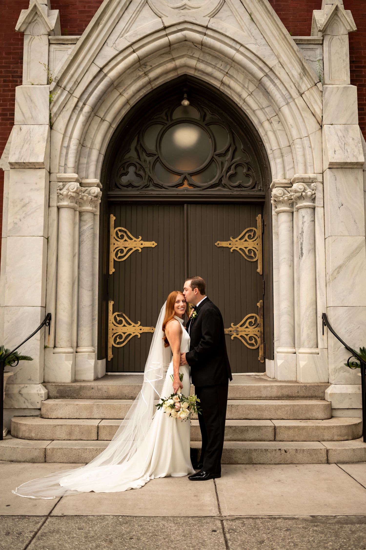 A couple poses in front of an ornate gothic church doorway with decorative bronze hinges and white stone archway.