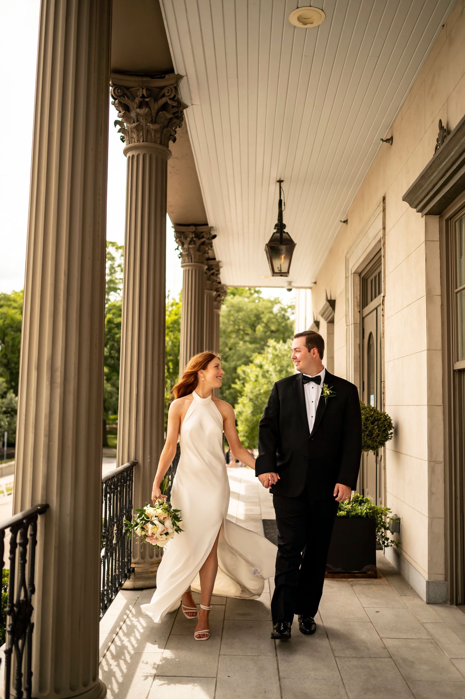 A couple in formal attire walk hand in hand along an elegant porch with classic columns and a hanging lantern.