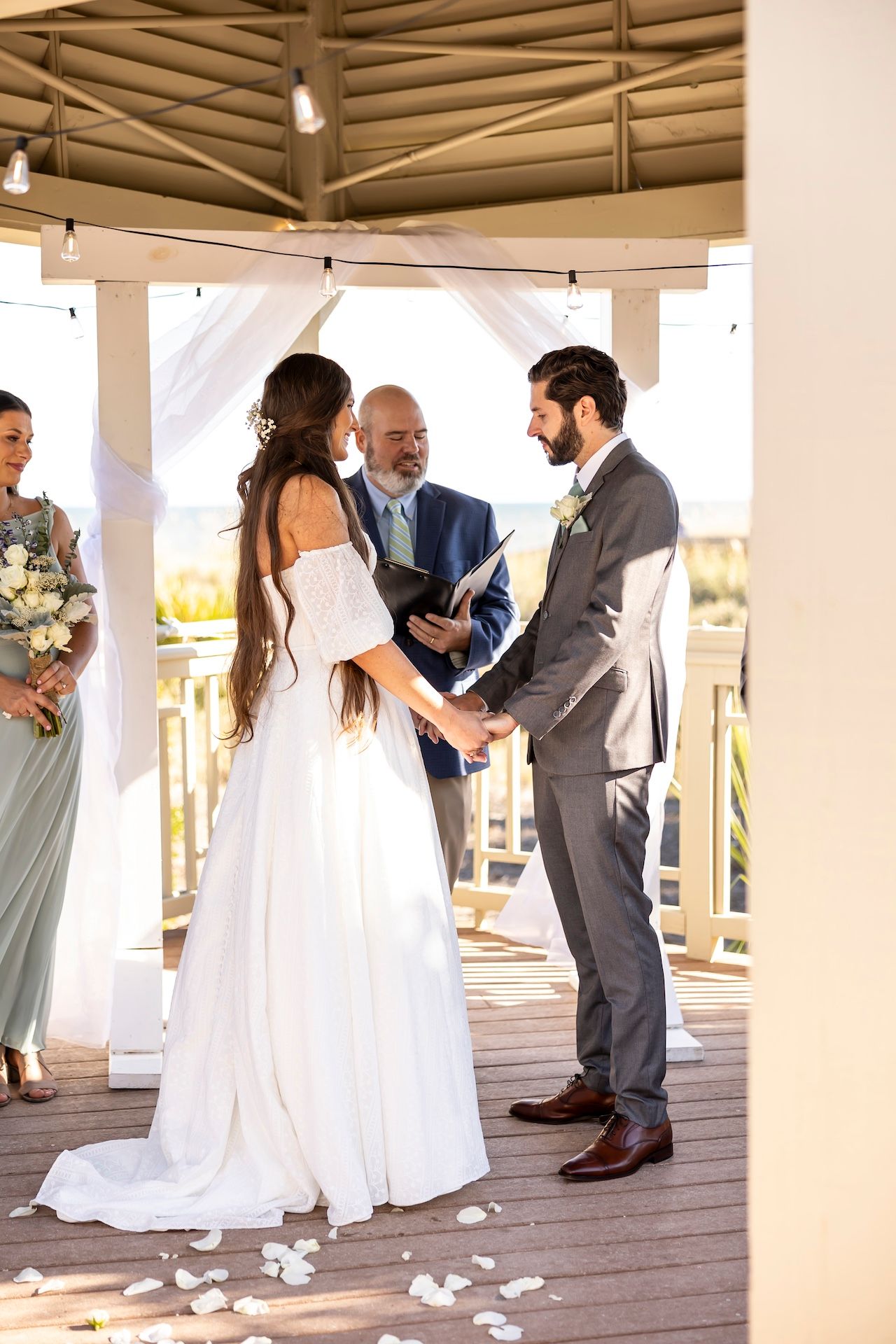 A wedding ceremony takes place on a covered deck with white drapery and flower petals scattered on the wooden floor.