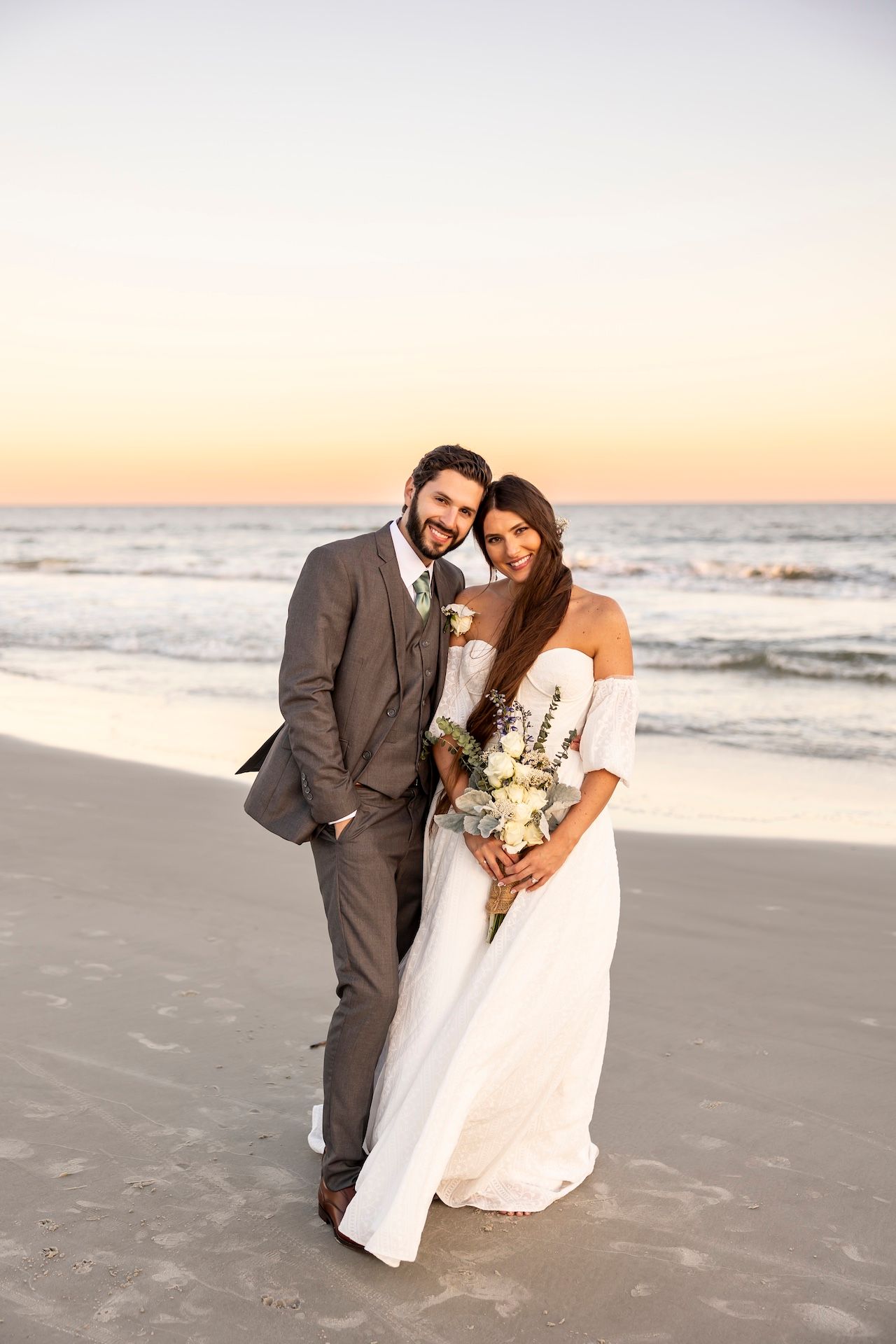 A couple poses together on a beach at sunset with waves crashing in the background and a white bridal bouquet in hand.