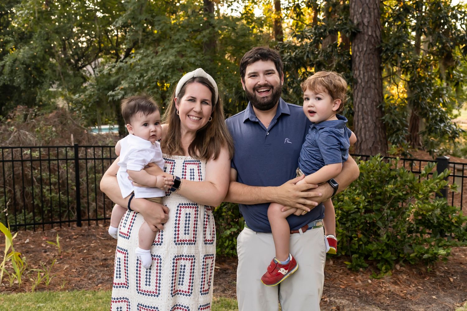 A family poses together outdoors in a backyard garden setting with trees and a black metal fence in the background.