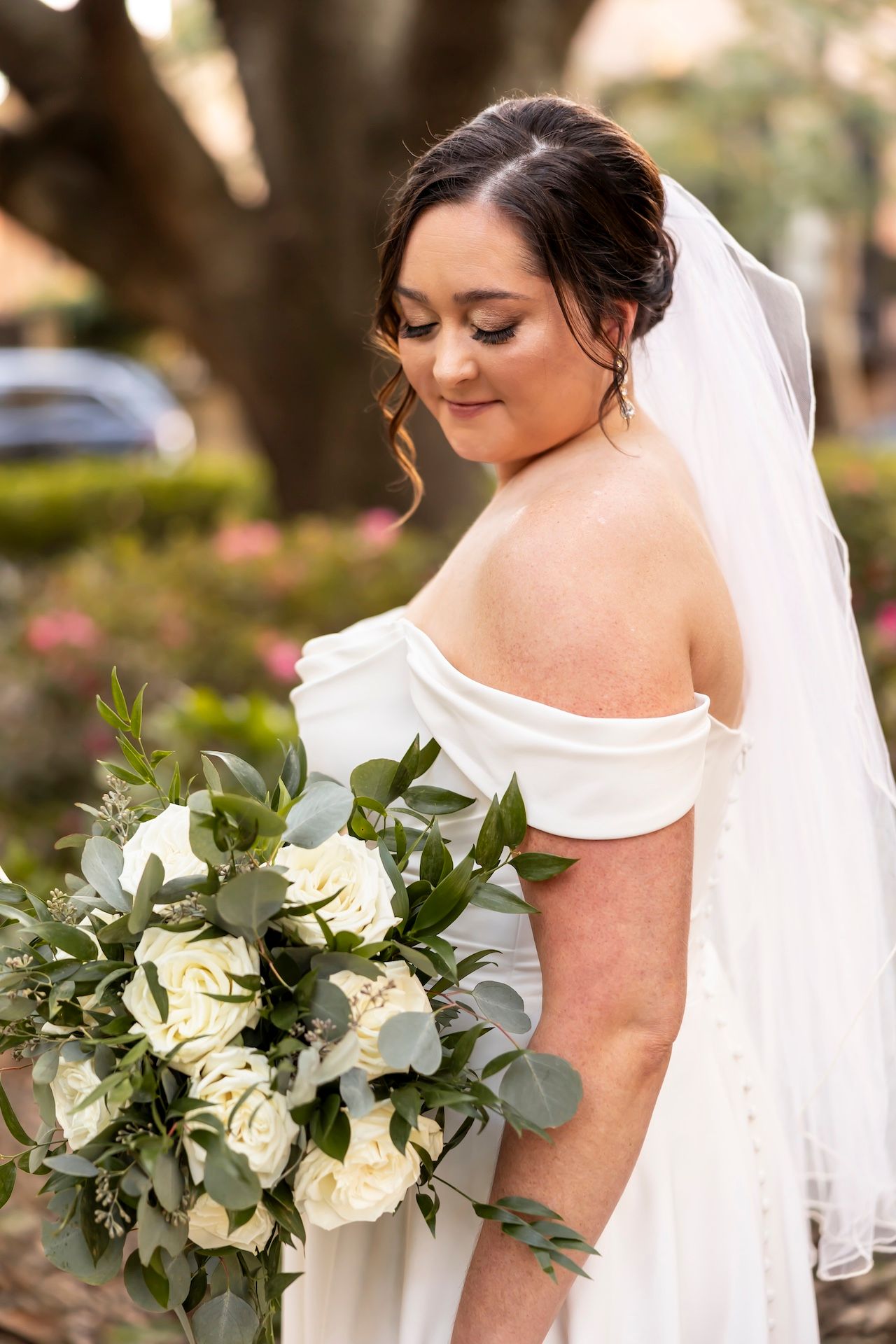 Bride in off-shoulder white dress holds elegant bouquet of white roses and eucalyptus leaves while looking down.