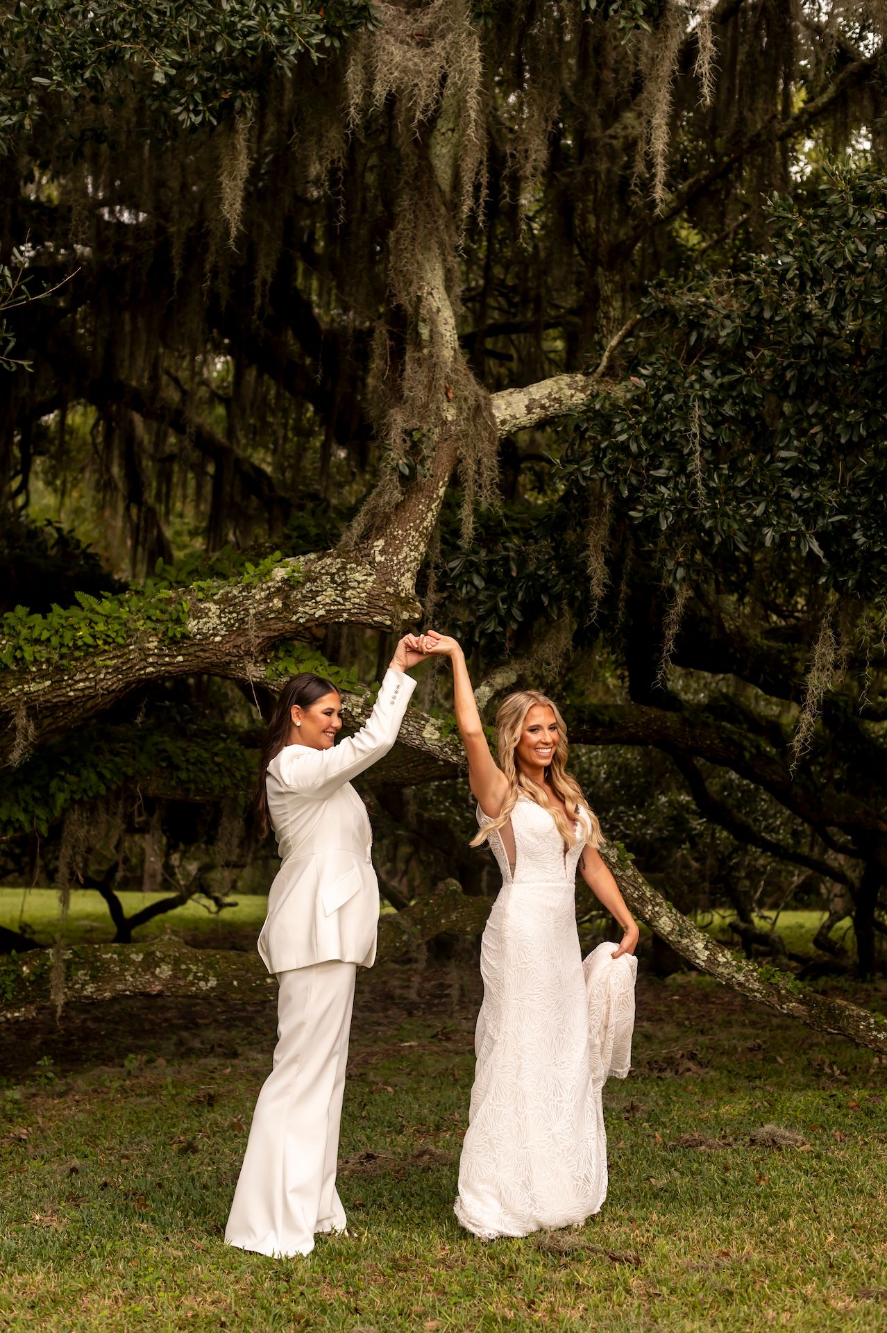 Couple in white outfits dances playfully together under moss-covered trees in an outdoor setting.
