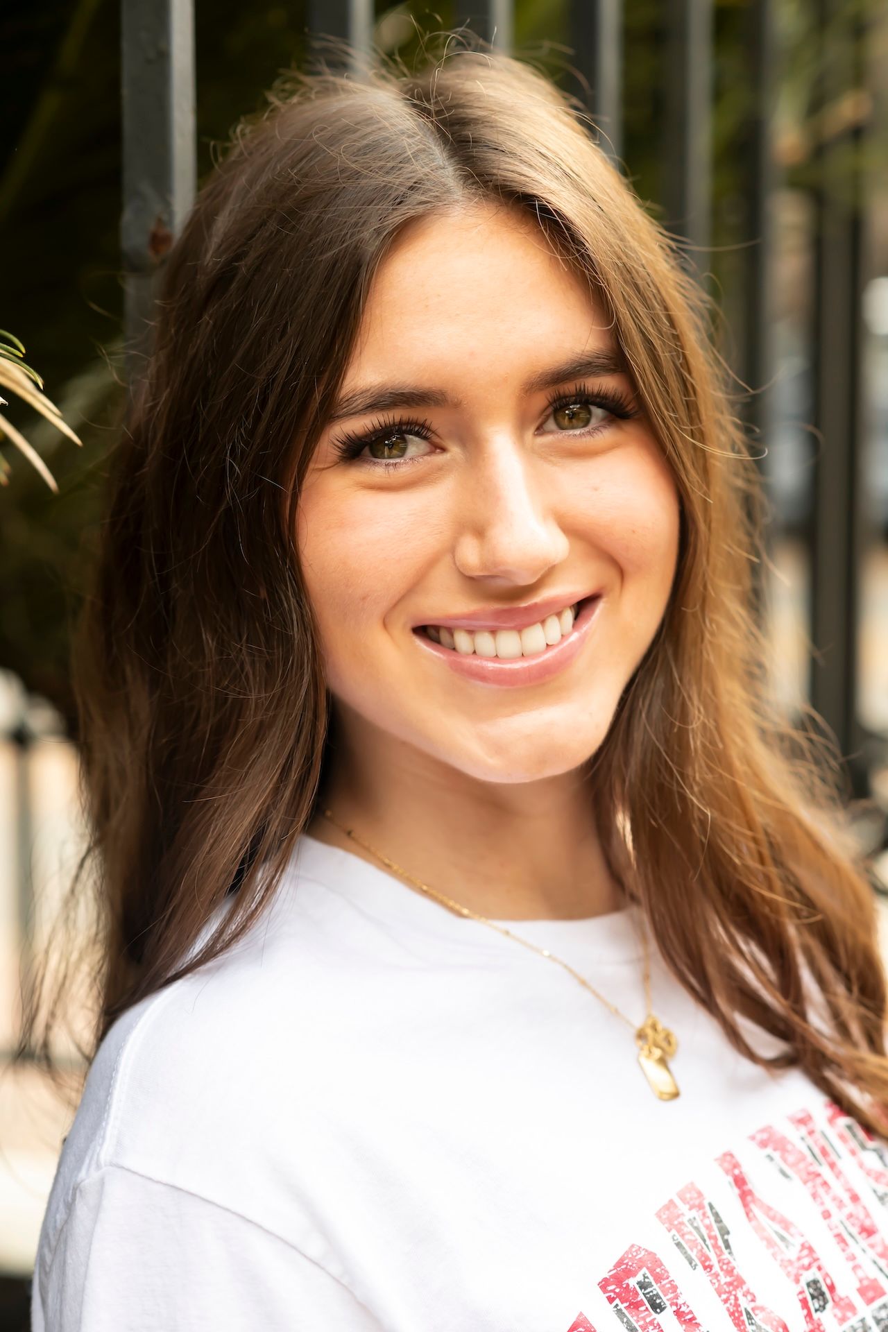 A person with long brown hair and a bright smile wearing a white t-shirt outdoors on a sunny day.