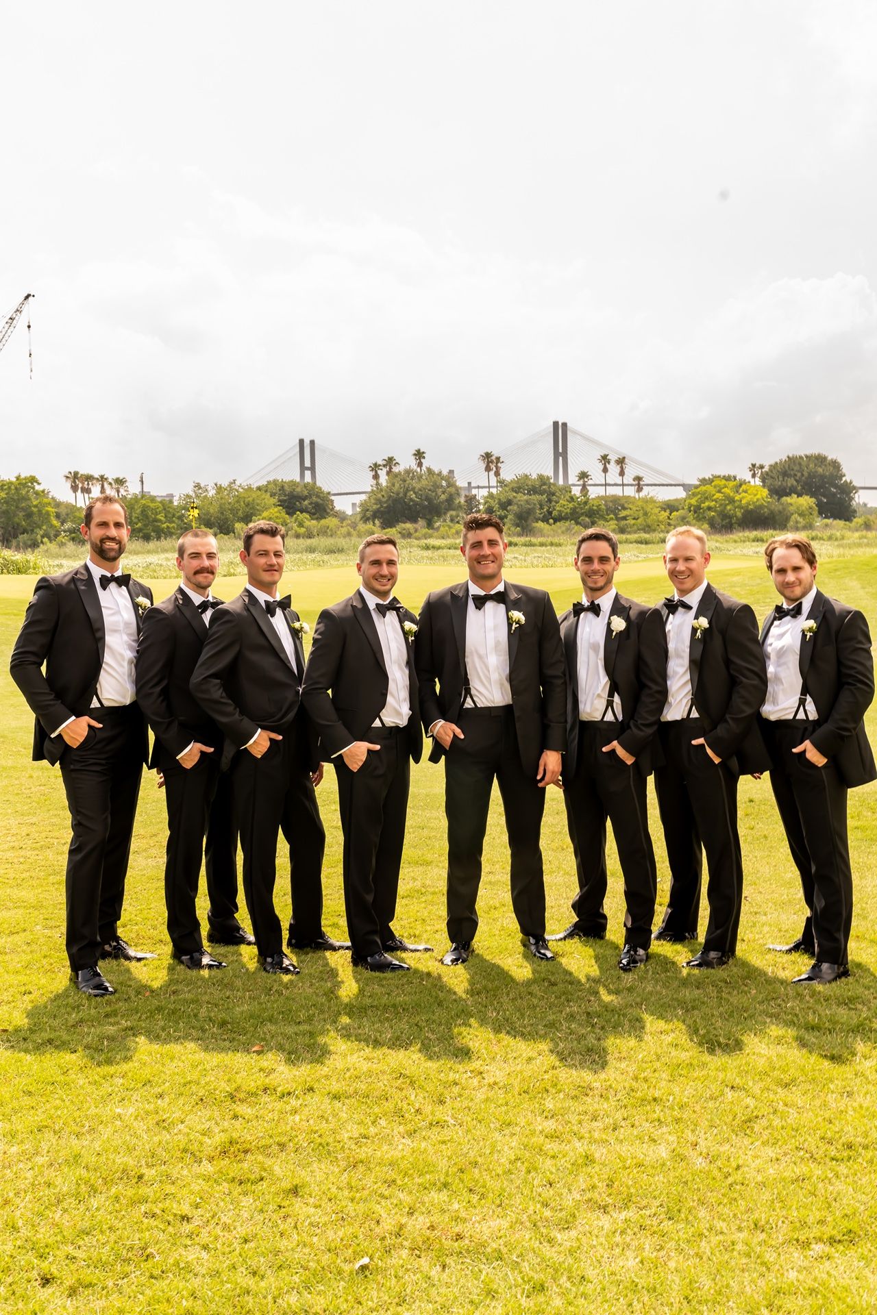 Groomsmen in black tuxedos pose together on grass field with industrial backdrop.