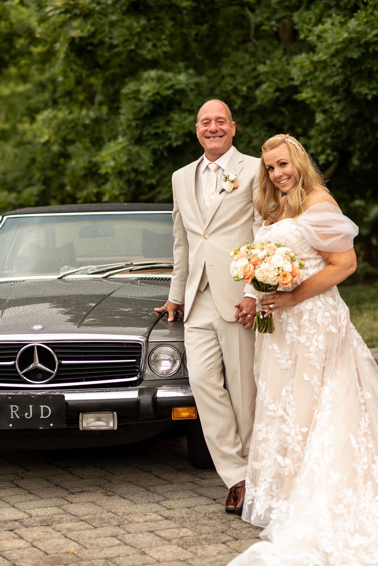 A newlywed couple poses beside a classic black Mercedes-Benz car, with the bride holding a peach and cream bouquet.