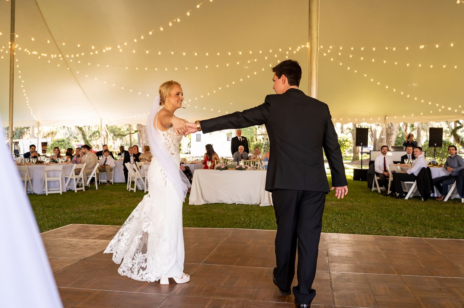A couple sharing their wedding dance on a tented dance floor with fairy lights above and guest tables in background.