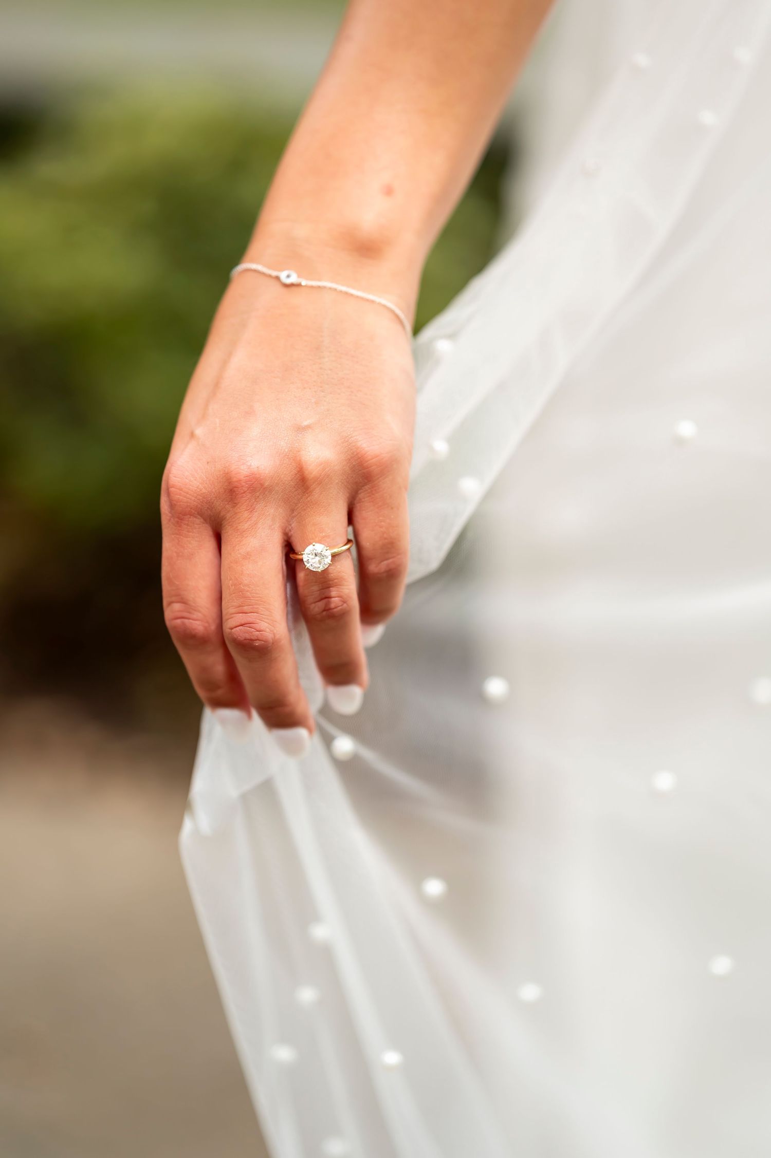 A delicate bride's hand wearing an engagement ring and bracelet while holding white tulle veil.