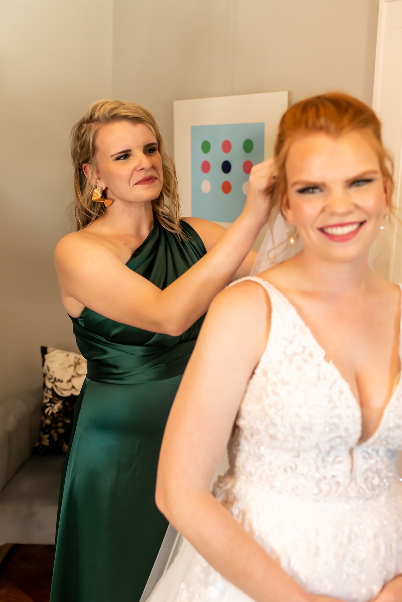 A woman in an emerald green formal dress helps adjust a bride's hair while getting ready in a white wedding gown.