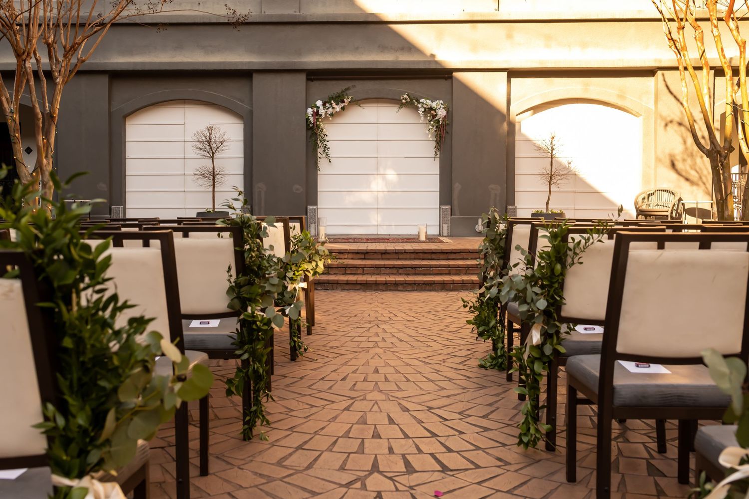 Indoor wedding ceremony venue with white chairs, brick pathway, and greenery decorations leading to an arched doorway.