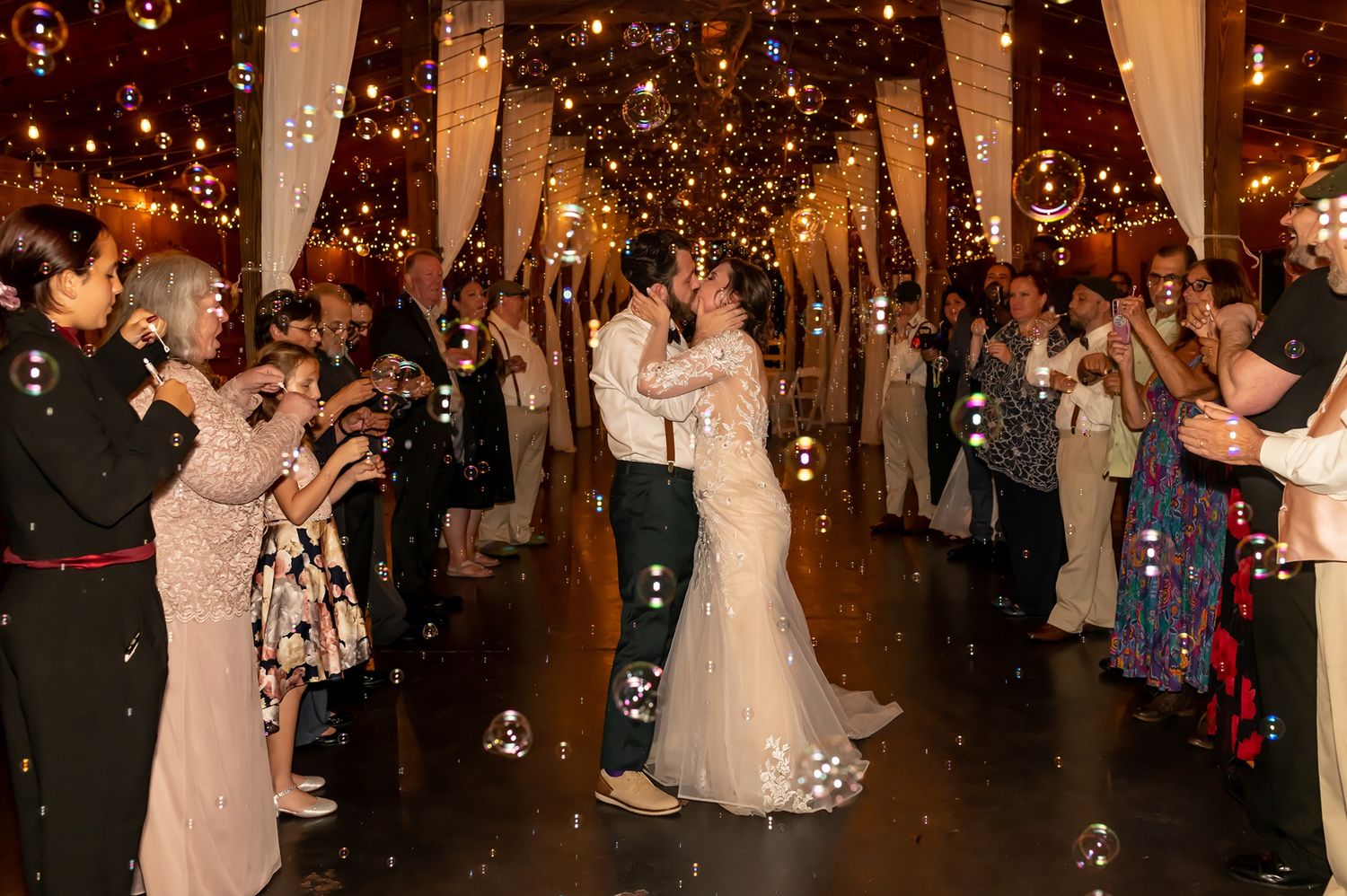 Couple shares romantic kiss during their first dance at wedding reception with guests and twinkling lights overhead.