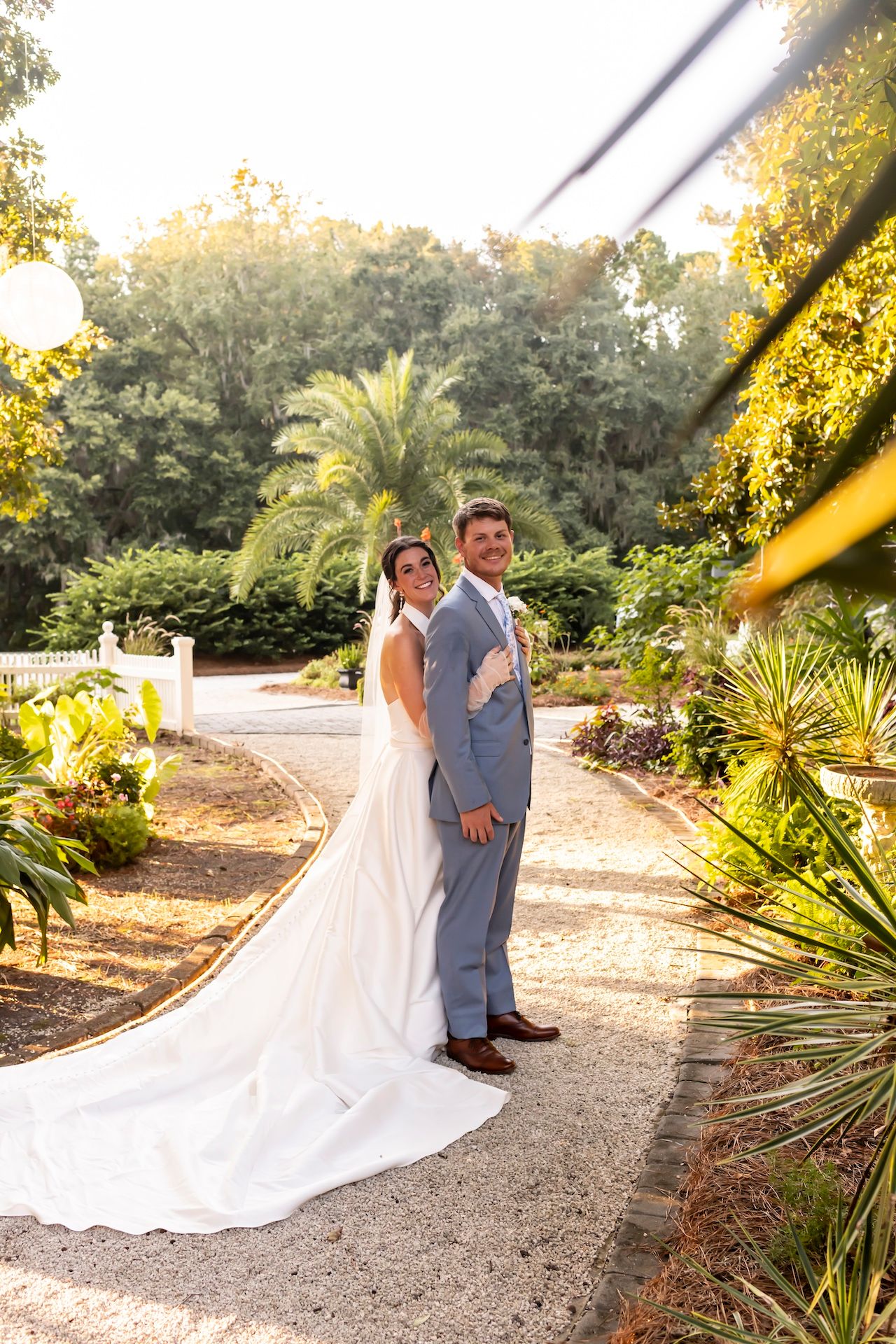 A couple poses in a lush garden setting with palm trees and foliage as golden sunlight filters through the trees.